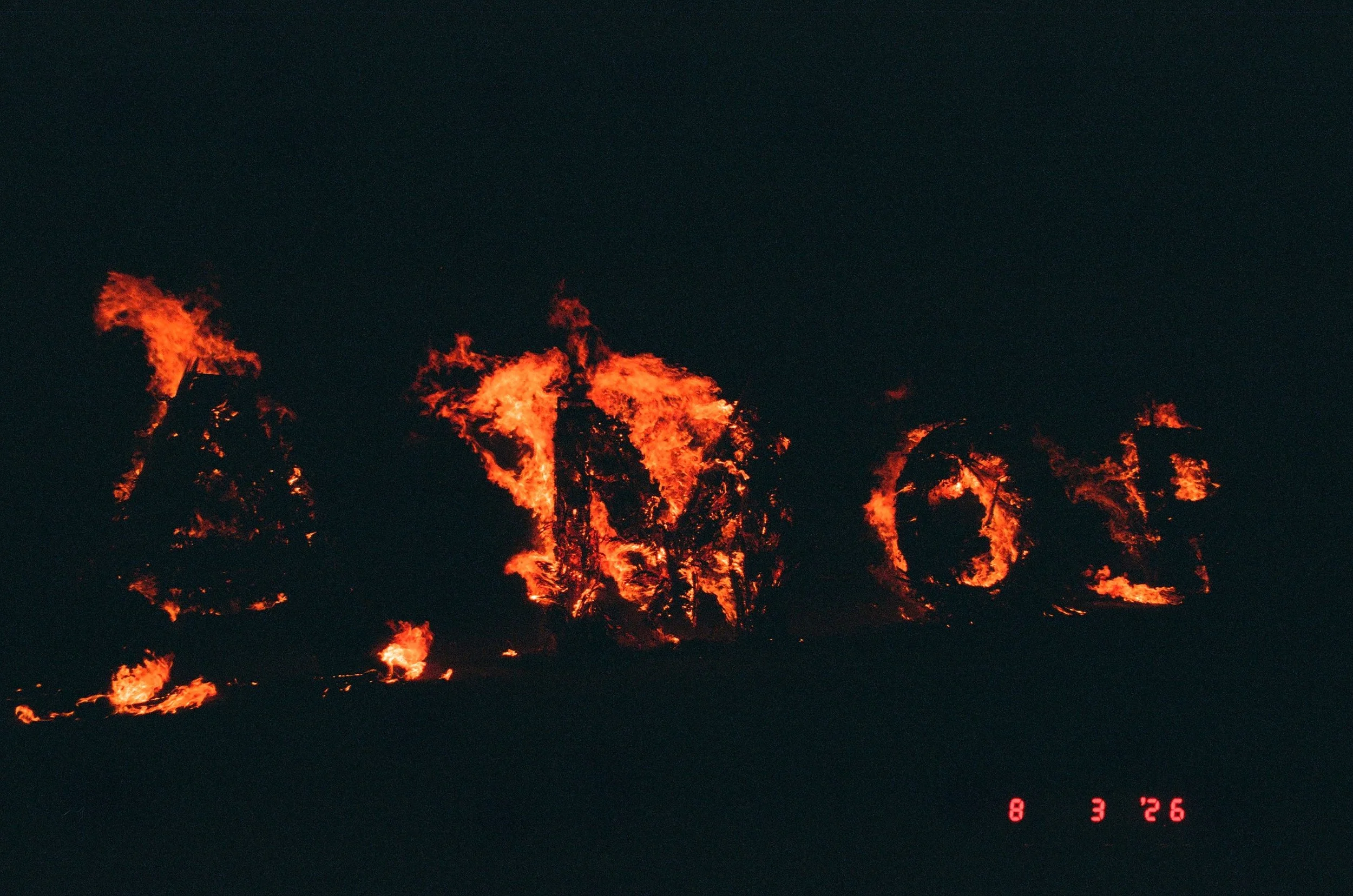 Nighttime fire burn on AMOR letters with visible flames and smoke, dark sky and red digital timestamp at bottom right.