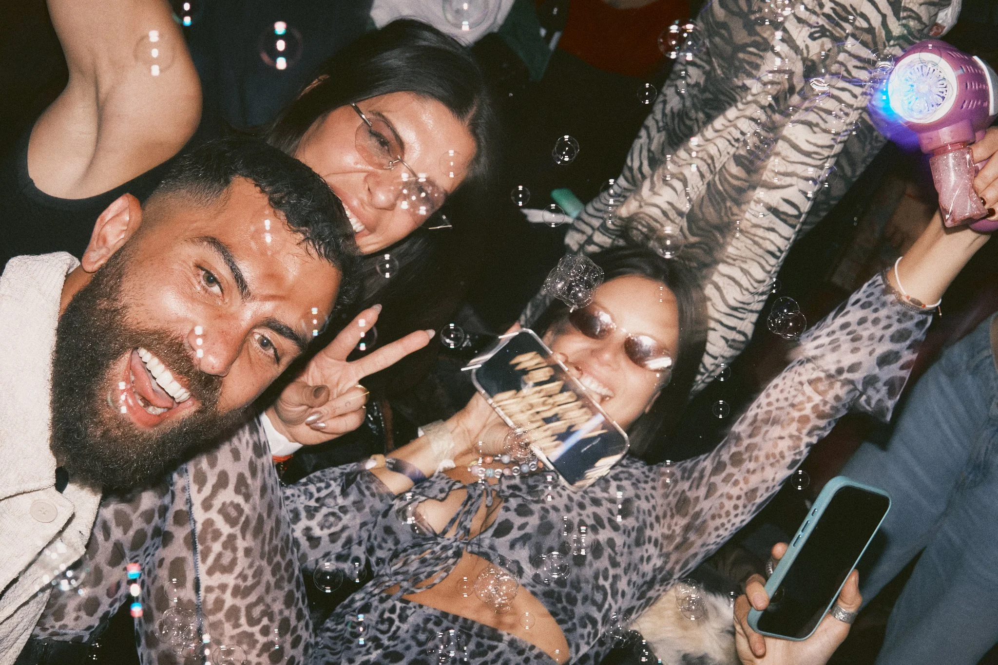A group of people celebrating at a party, taking a selfie with a smartphone, surrounded by bubbles, wearing animal print outfits, smiling, and making peace signs.