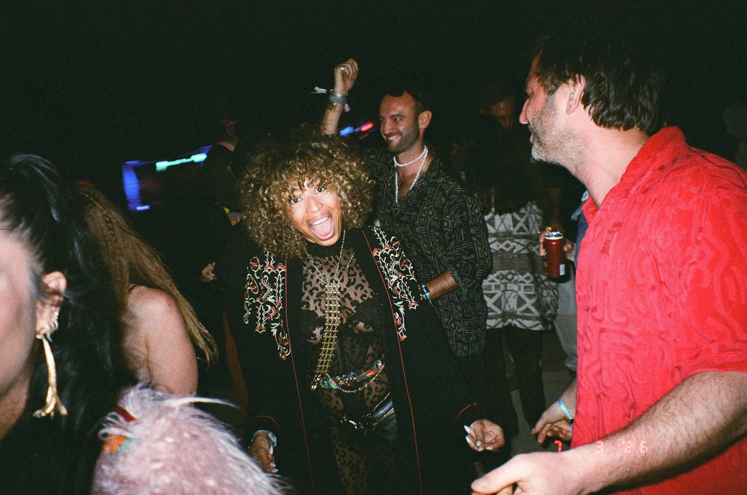 People dancing and socializing at Amarás Oaxaca Experience 2026, with a woman with curly hair smiling and wearing a patterned jacket and a man with a beard and a boutonniere in the background.