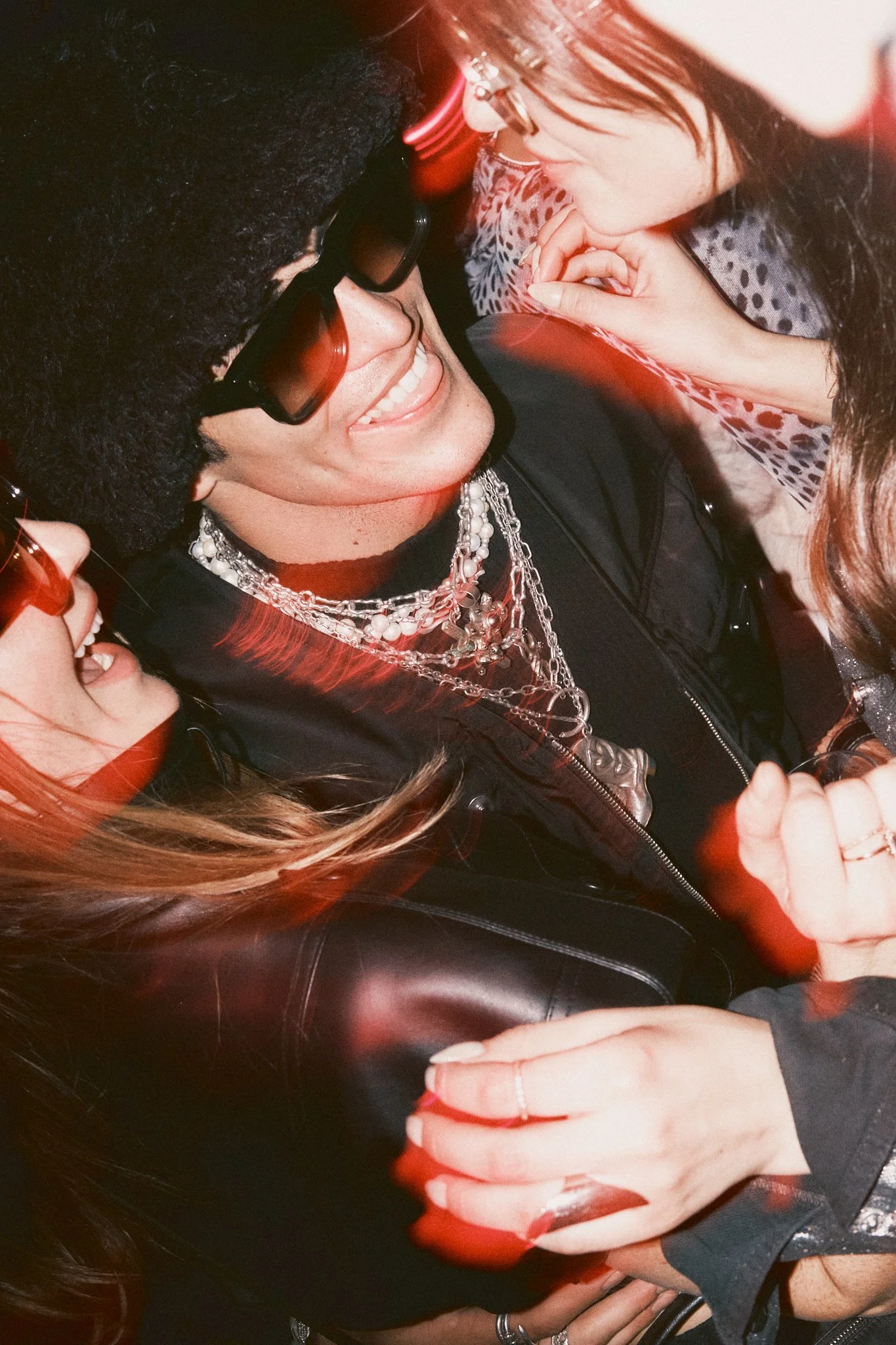 Group of people close together, smiling and engaging in a fun, lively moment, wearing fashionable accessories and jackets, with some wearing sunglasses and jewelry.
