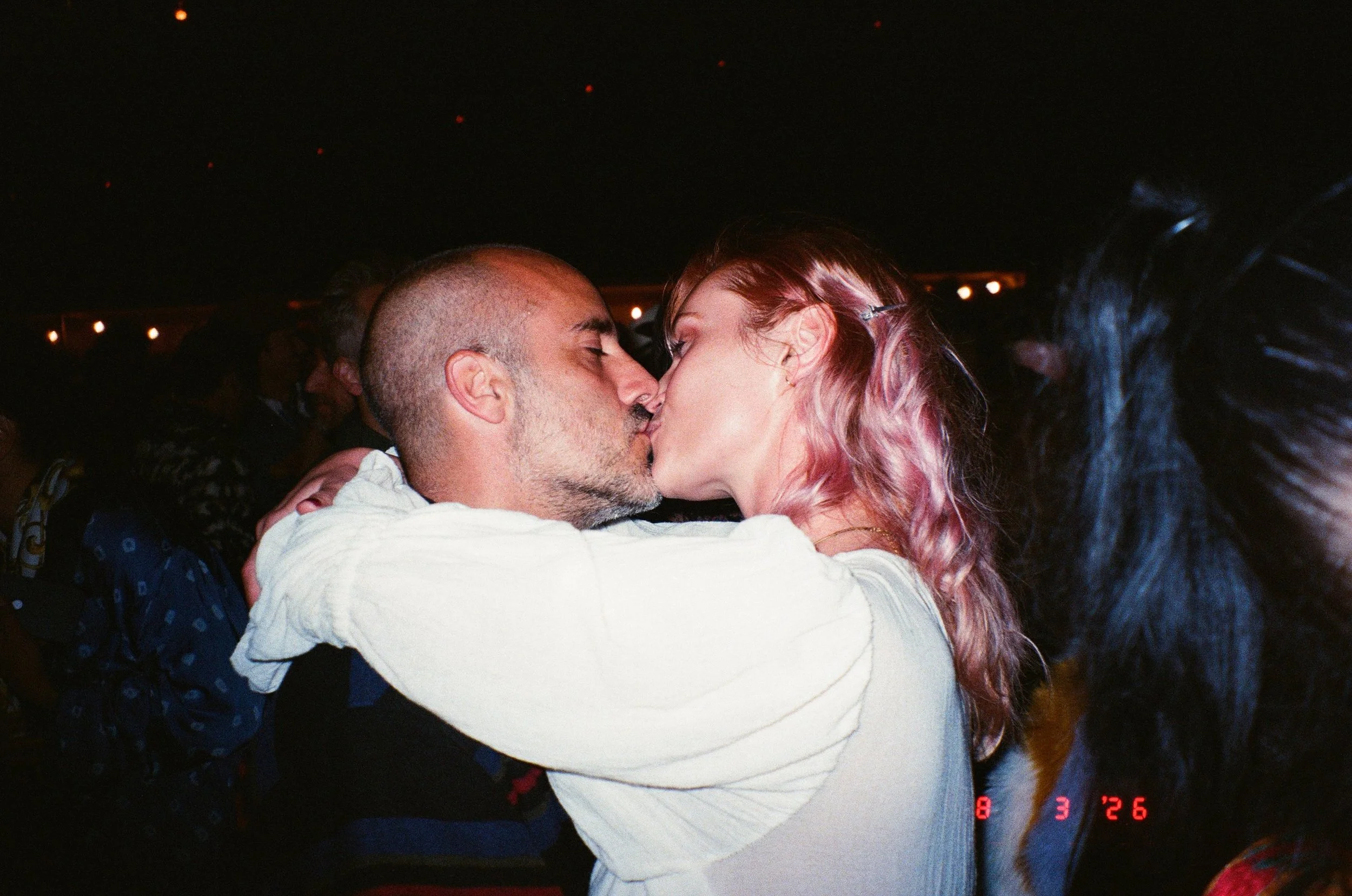 A couple kissing closely in a dark setting, with the woman having red and pink hair and the man with a shaved head and beard at Amarás Oaxaca Experience 2026.