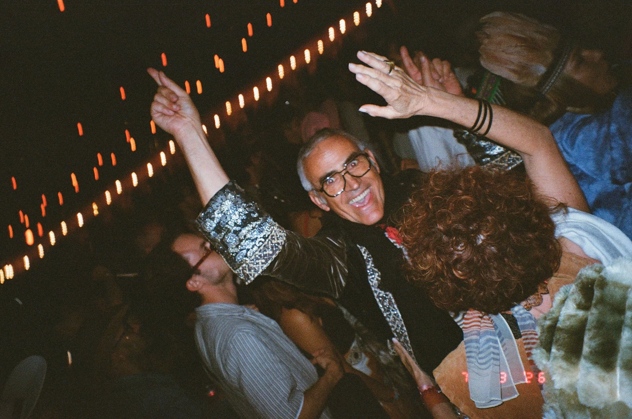 A group of mature people dancing and celebrating at Amarás Oaxaca Experience 2026, with one woman in the center smiling widely and wearing glasses, surrounded by others raising their hands and enjoying the moment. String lights are visible overhead.