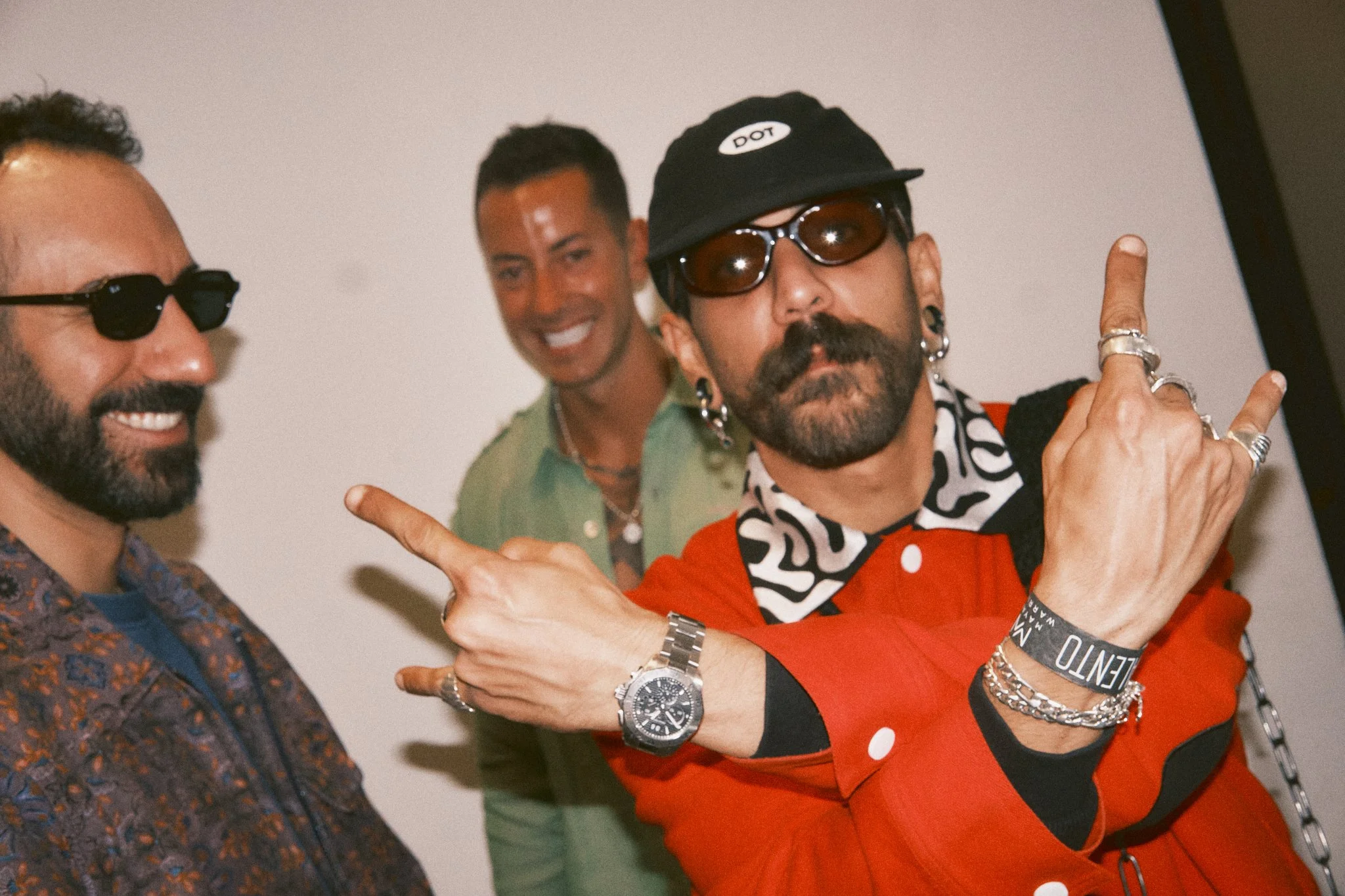 Three men smiling and posing, one wearing sunglasses and a patterned shirt, another in a green shirt, and OMRI. in an orange jacket with accessories, sunglasses, and a cap, in the green room.
