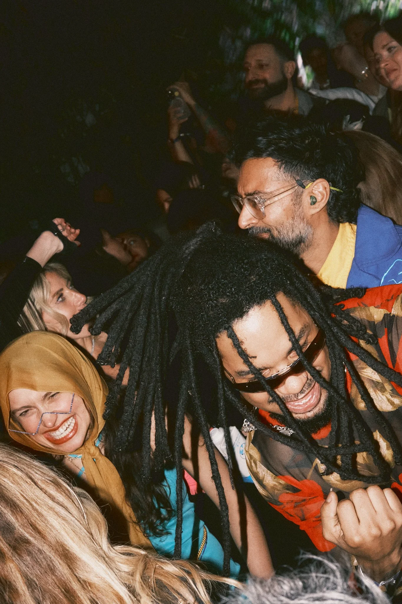 A lively crowd at a party or concert, with people dancing and enjoying themselves. The focus is on a man with long dreadlocks and sunglasses, smiling and showing his bicep, surrounded by smiling women.