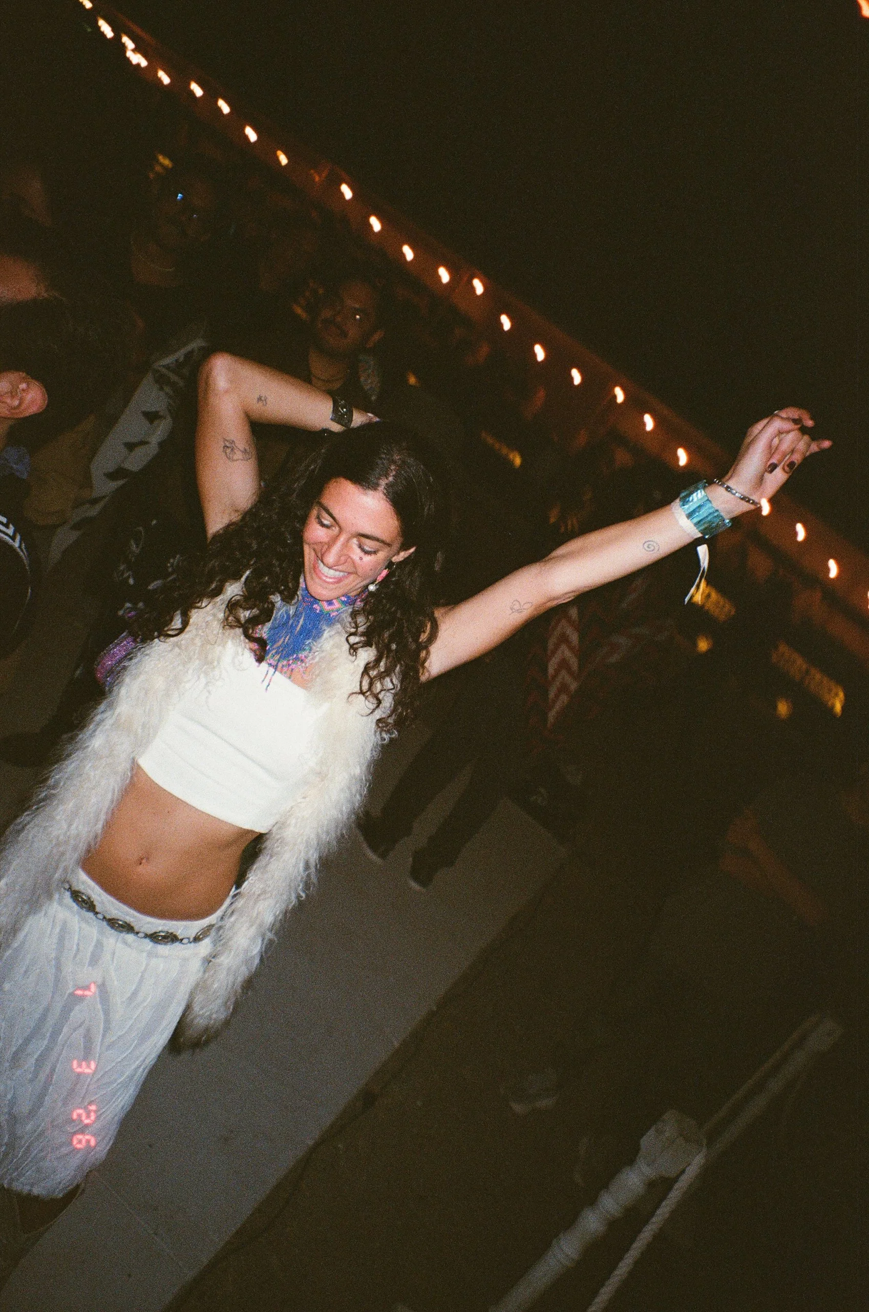 A woman with curly hair singing and dancing at Amarás Oaxaca Experience 2026, wearing a white furry vest and a matching skirt with her arms raised.