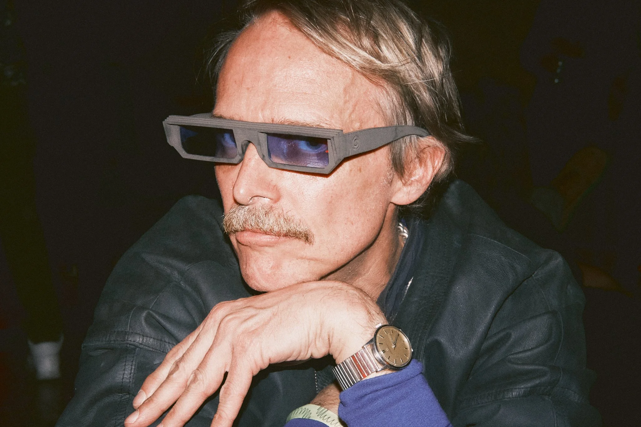 A man with a mustache and gray hair wearing Octo Ocular eyewear glasses, a black leather jacket, and a silver watch on his left wrist, resting his chin on his hand.