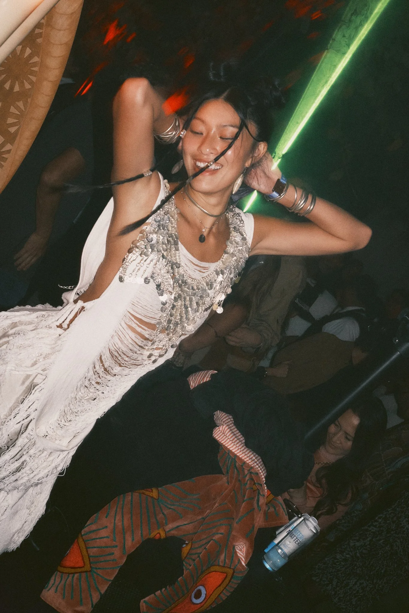 A woman dancing in a club, wearing a white fringed dress with silver embellishments, multiple necklaces, and bracelets, with her eyes closed and a joyful expression.