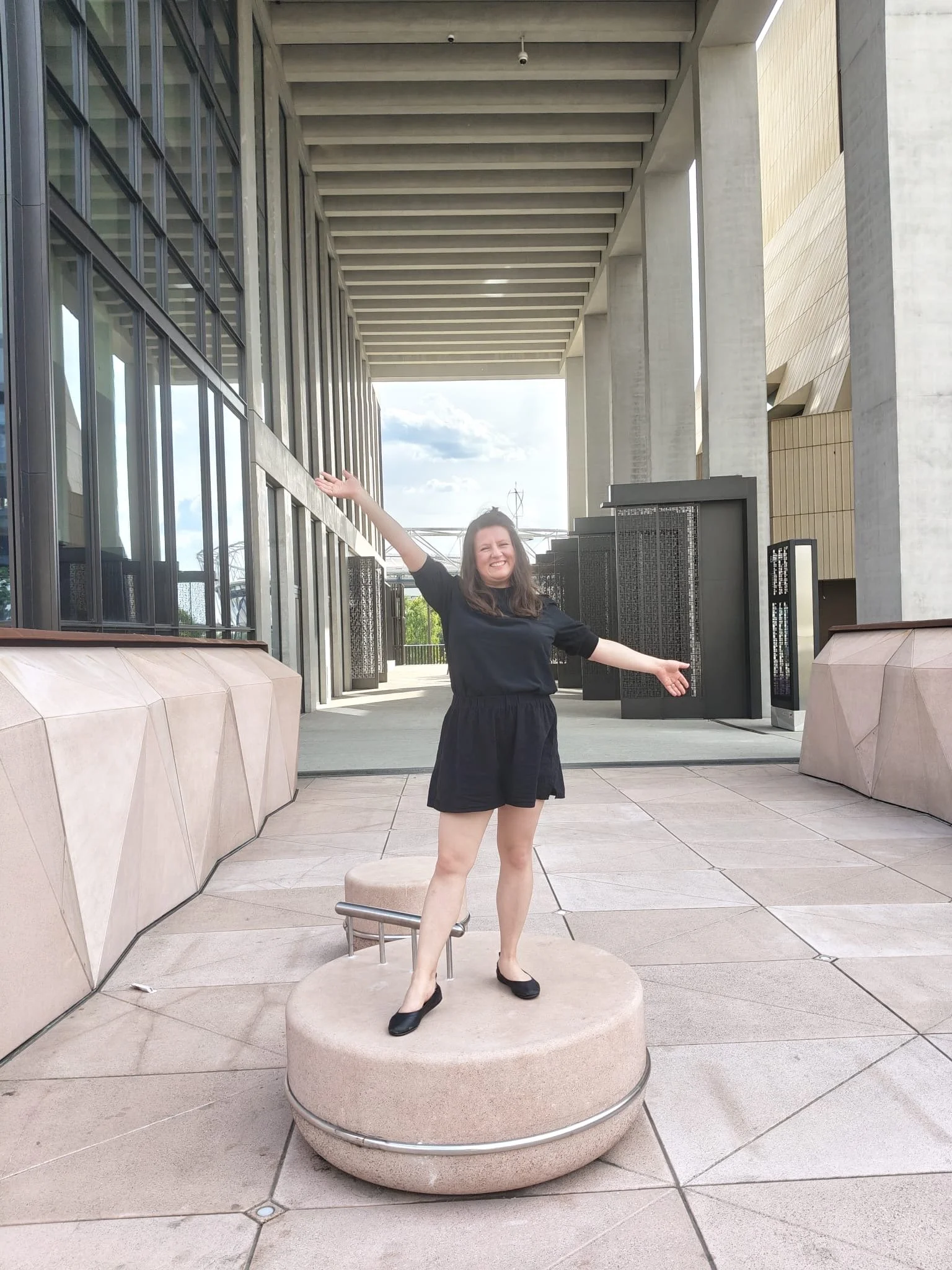 Picture showing Ania Feranska-Iqbal, Speaking Voice Coach and Founder of Voice Stories London. Ania is standing on a circular column with her arms open and spread out to showcase how much space there is around her. She's smiling.
