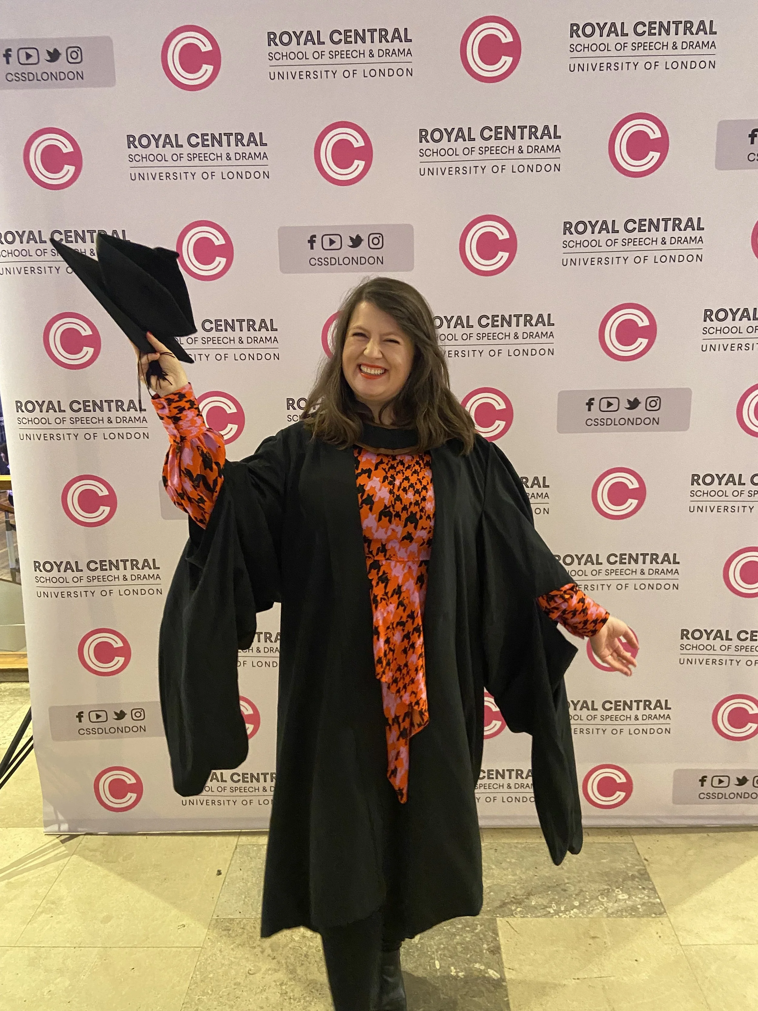 Ania Feranska-Iqbal on her graduation day smiling and happily posing in front of the Royal Central School of Speech and Drama banner.Ania is holding her graduation hat up in her right hand.