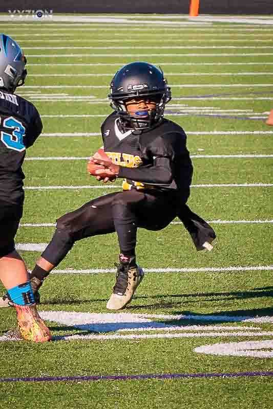 Young football player in black uniform and helmet crouching on a football field, holding a football in both hands, preparing to run or pass.