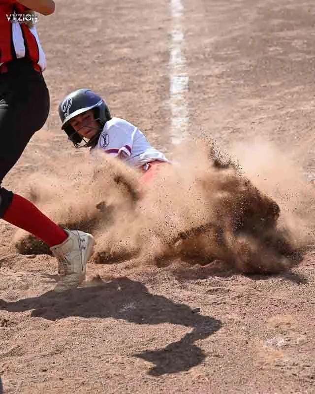A young girl sliding into a base during a softball game, with a dust cloud around her, and an adult standing nearby.