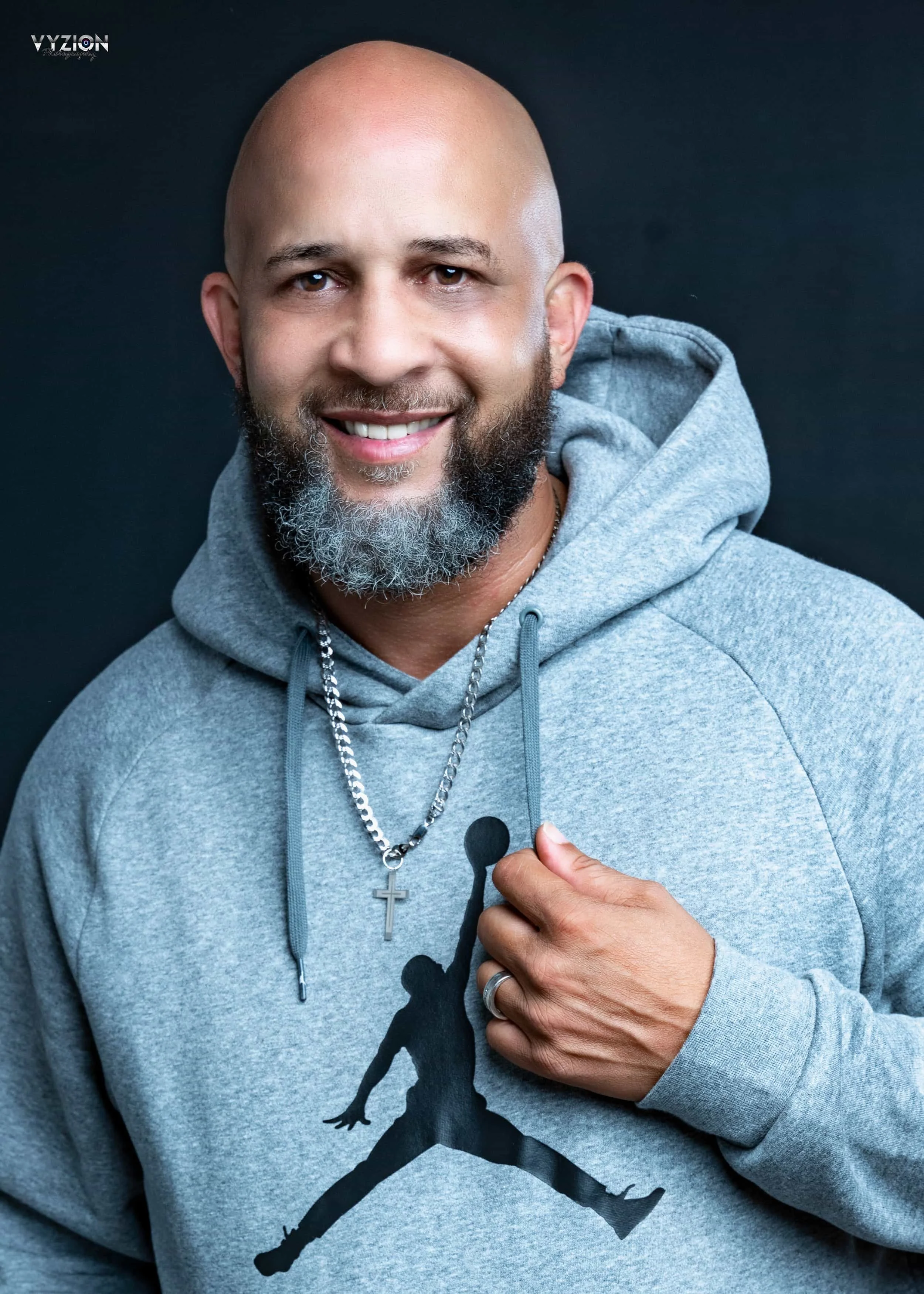 Smiling man in gray hoodie with basketball silhouette logo, wearing a cross necklace.