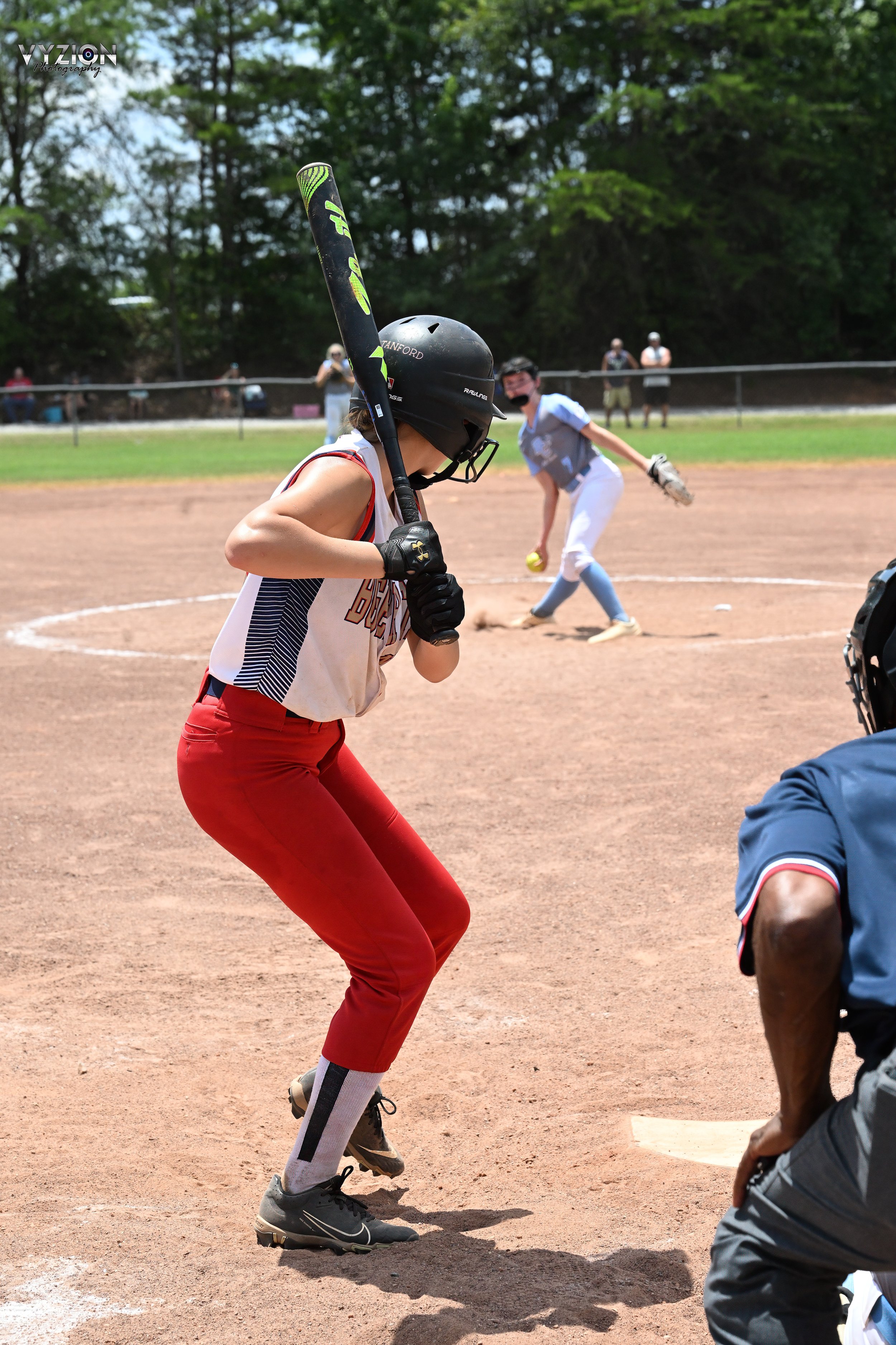 Youth softball player at bat in game, with pitcher mid-throw on sunny day.