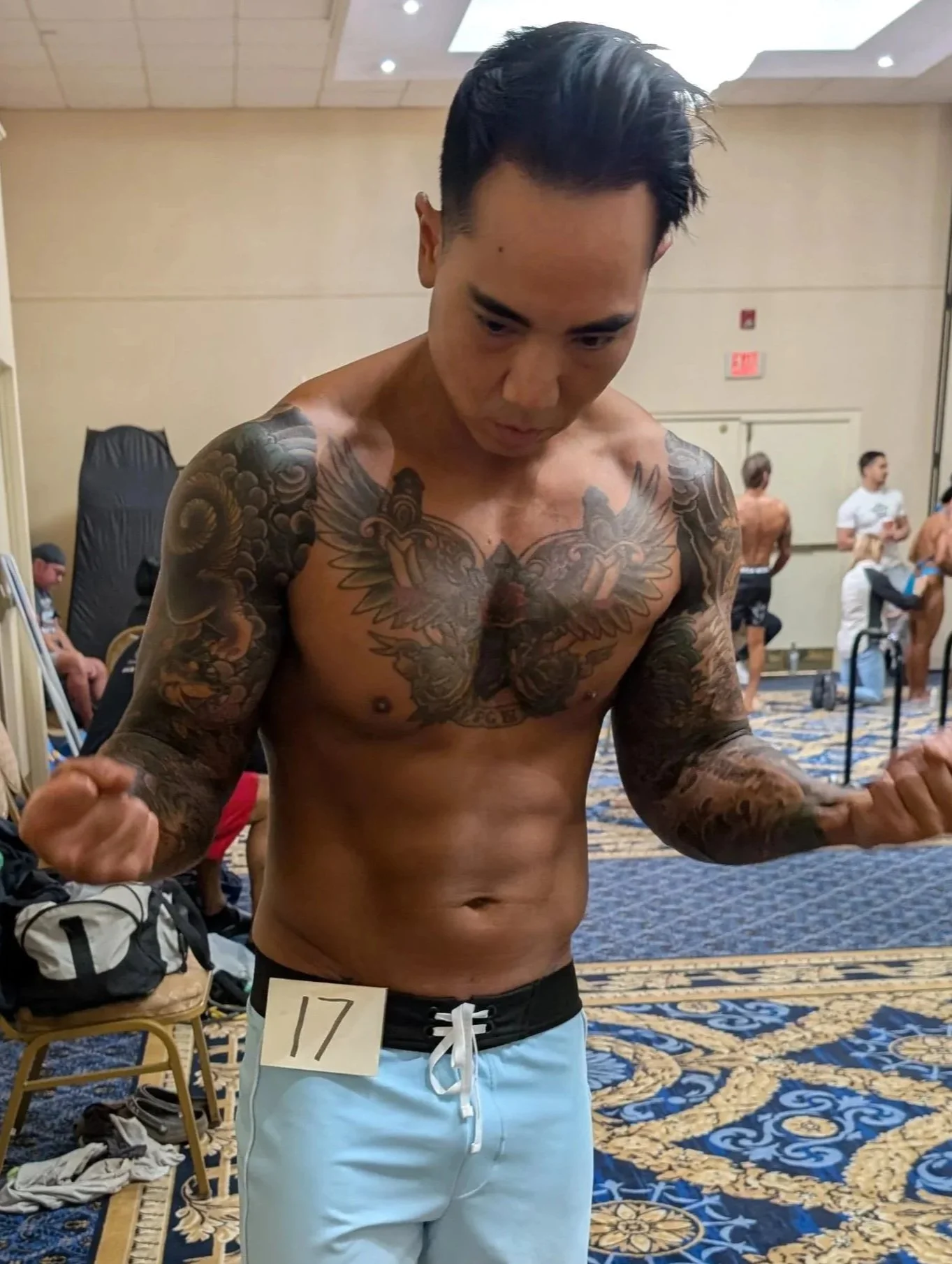 A shirtless man with tattoos on his chest and arms, standing in a room with a patterned carpet, holding his fists up in a fighting pose, with a number 17 on his waistband.