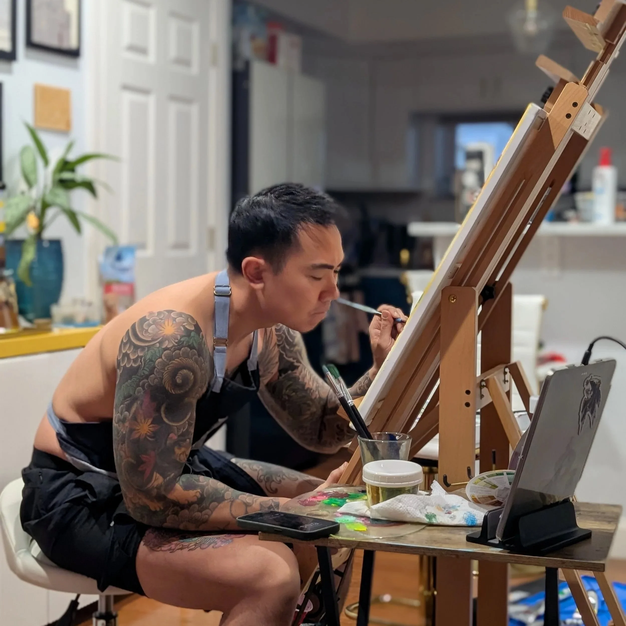A man with tattoos painting at an easel in a kitchen, sitting on a stool, wearing only shorts and an apron.