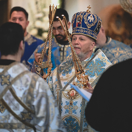 Byzantine bishop wearing a richly decorated blue and gold mitre and vestments, holding ornate gold candleholders during a religious ceremony with other clergy and congregation members in similar vestments.