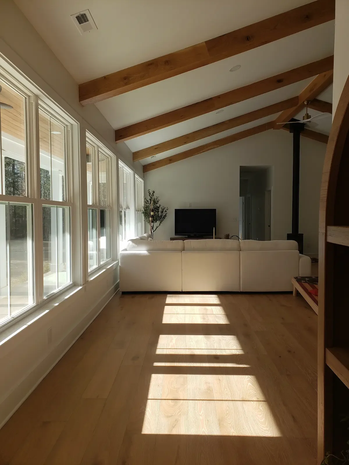 Living room with large windows on the left, a white sectional sofa facing a black TV, exposed wooden beams on the ceiling, a ceiling fan, and a small indoor plant near the TV.