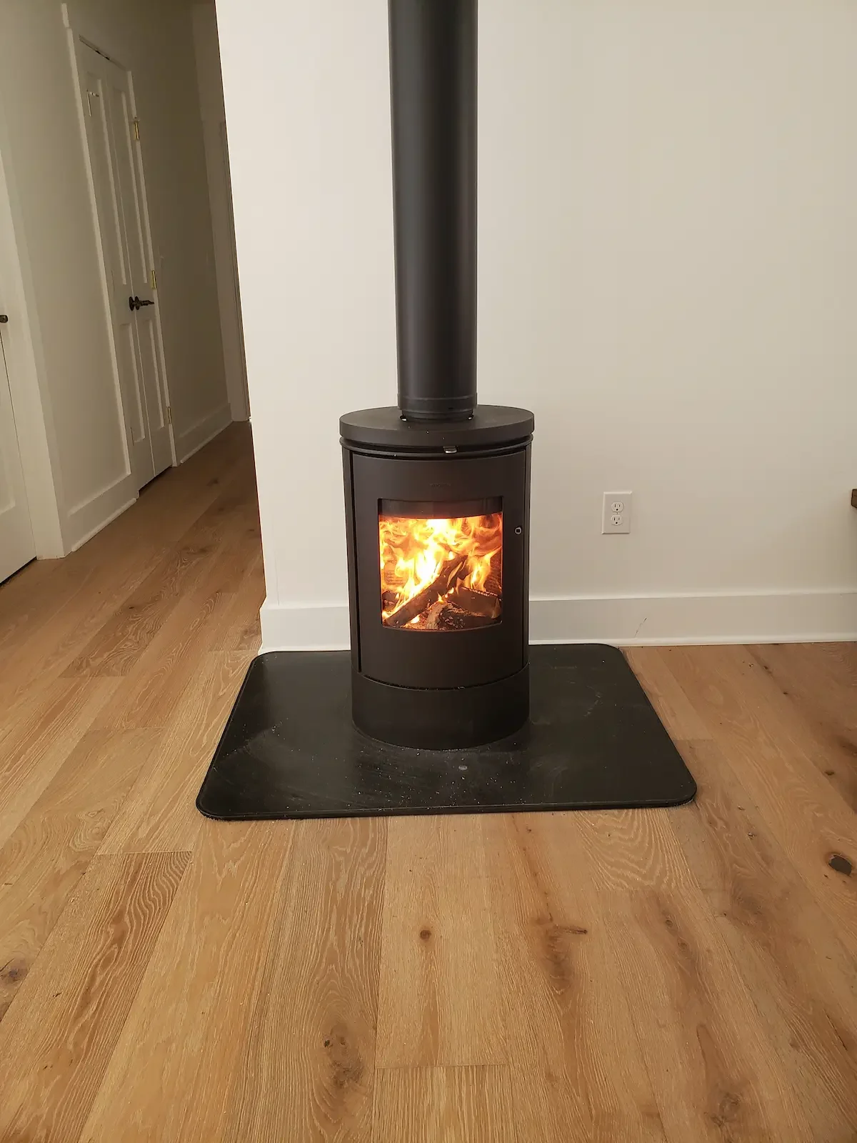 Black wood-burning stove with a glass door showing flames inside, on a black mat in a room with wooden floor and white walls.