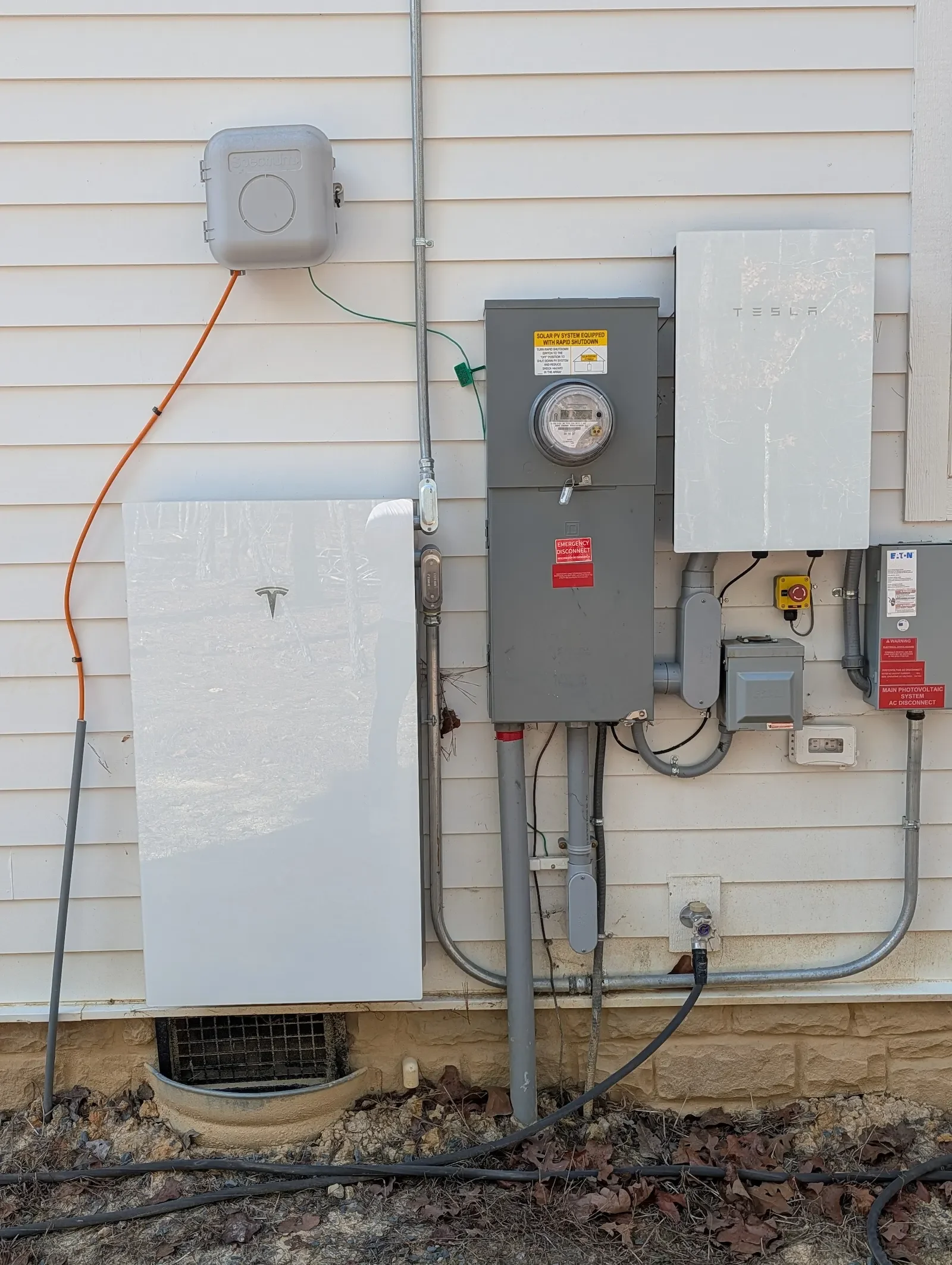 Exterior view of a house showing electrical and solar power system components including a Tesla wall, electrical boxes, and wiring both on the wall and the ground.