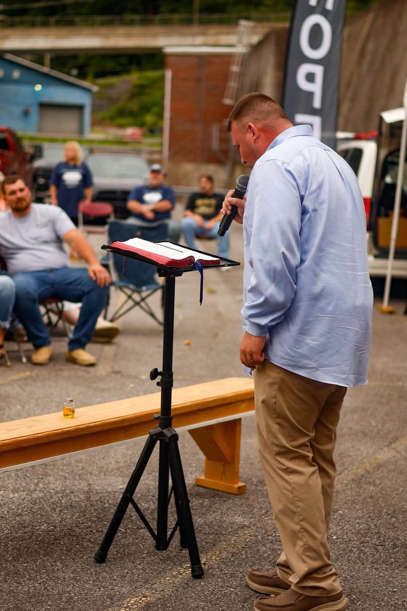 Man in a light blue shirt and khaki pants speaking into a microphone while reading from a clipboard at an outdoor event. Several people seated in the background, some with arms crossed, watching him. A small bottle of honey on a wooden bench in front of him, and a large vertical black banner with white text partially visible. The setting appears to be a parking lot with vehicles and buildings in the background.