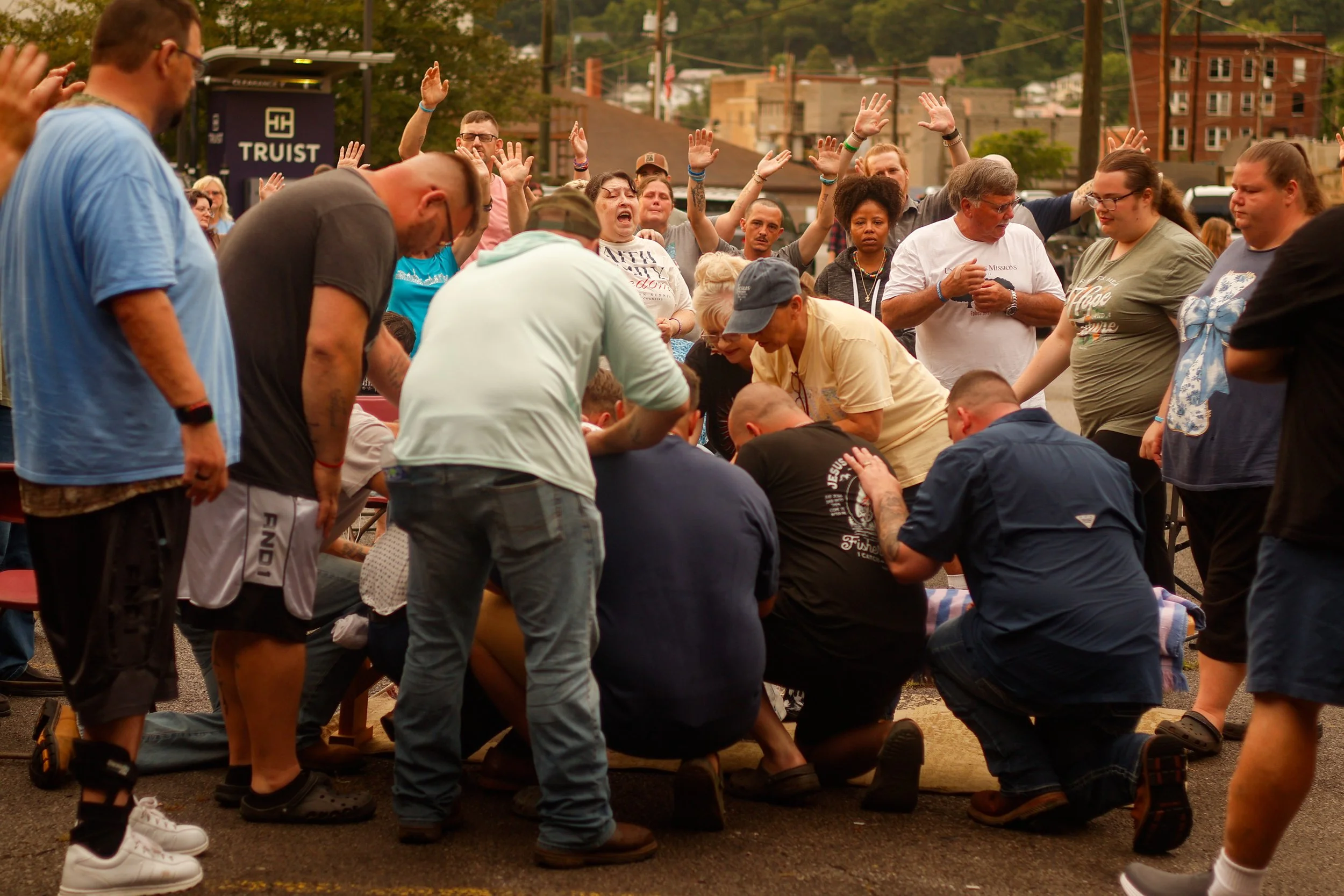 A group of people participating in a prayer or religious gathering outdoors, with hands raised and heads bowed, standing around a person kneeling on the ground.