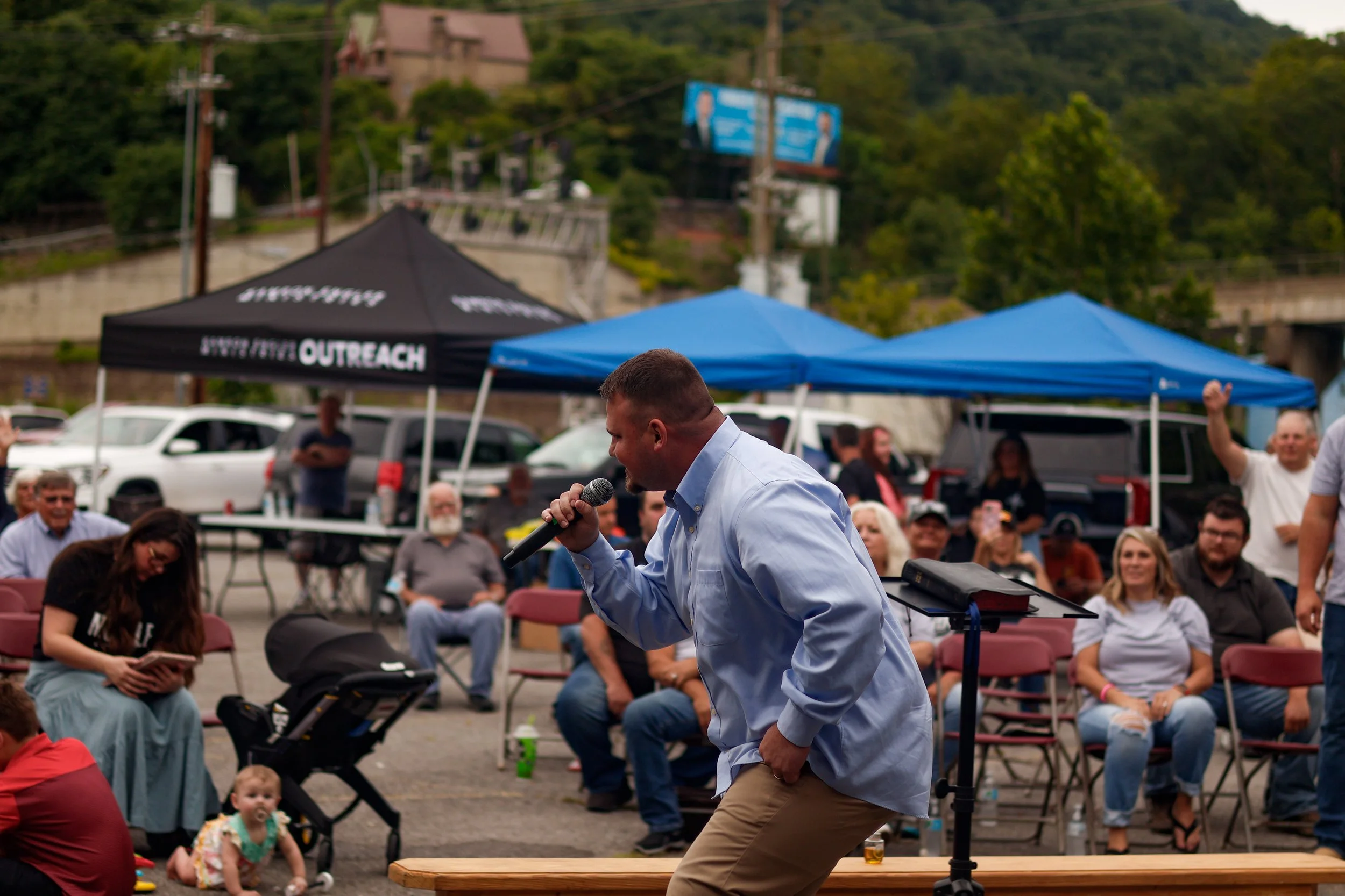 A man in a light blue shirt and khaki pants speaking into a microphone at an outdoor event, with an audience seated behind him under tents, including a woman with a stroller and a baby, and a man raising his fist in applause. There are trees, cars, and a hillside with buildings in the background.