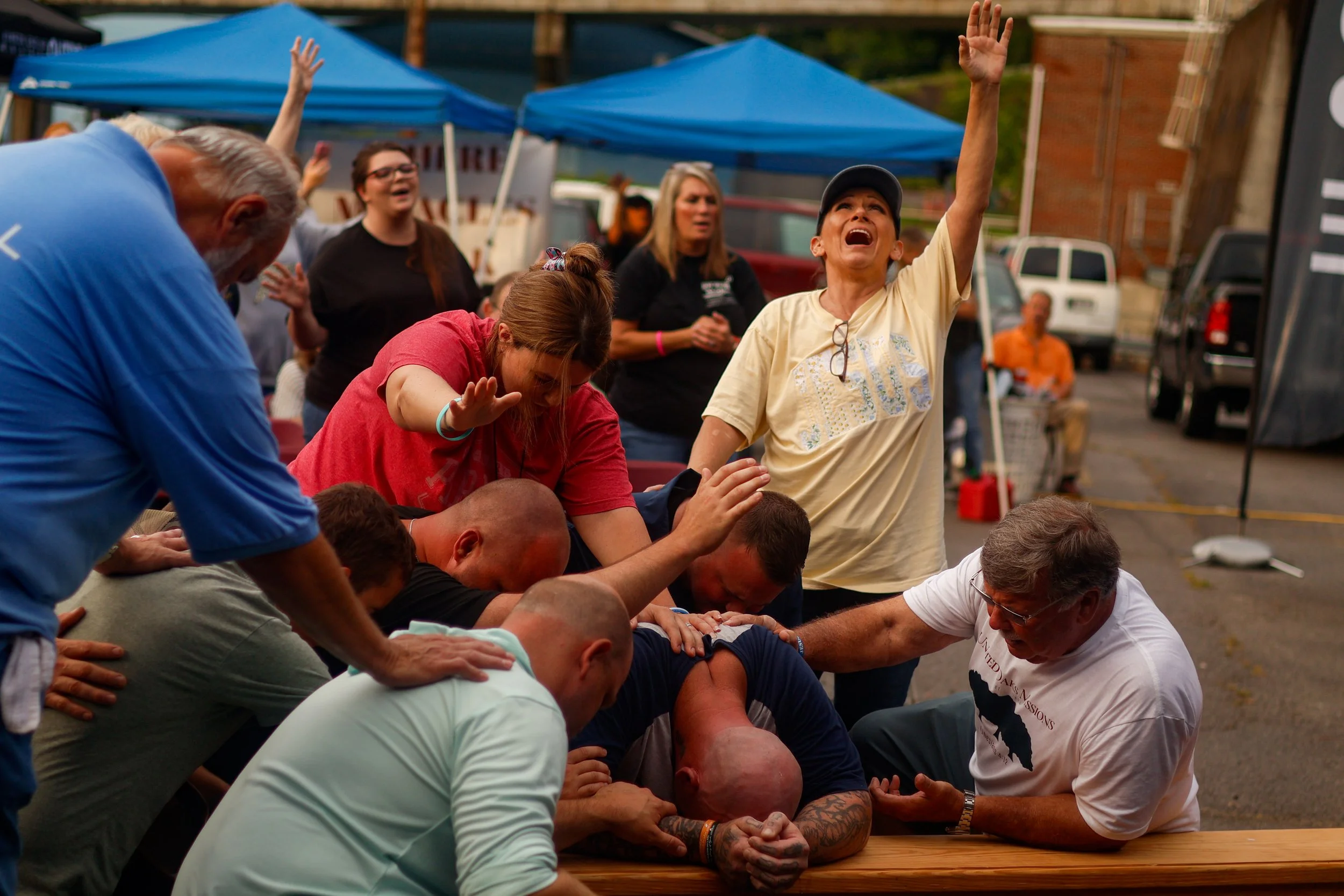 People participating in a door brawl, their arms intertwined, with a woman looking upwards and others appearing distressed or engaged in the activity, outdoors under blue tents.