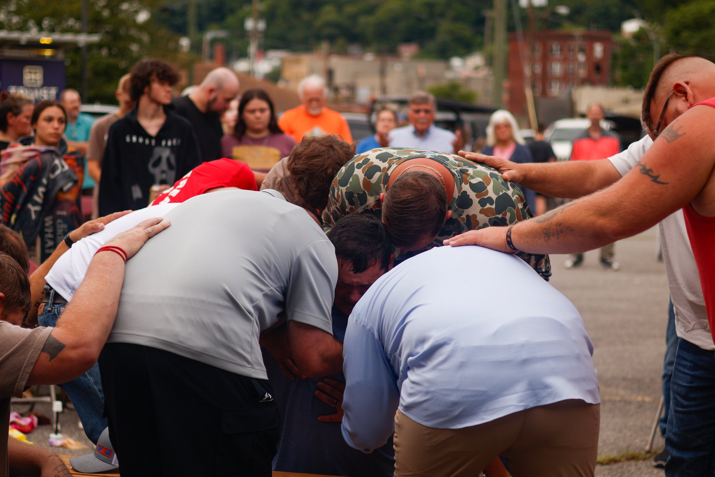 A group of people, including men and women, participate in an outdoor prayer circle, bending over with hands on each other's shoulders, with onlookers in the background