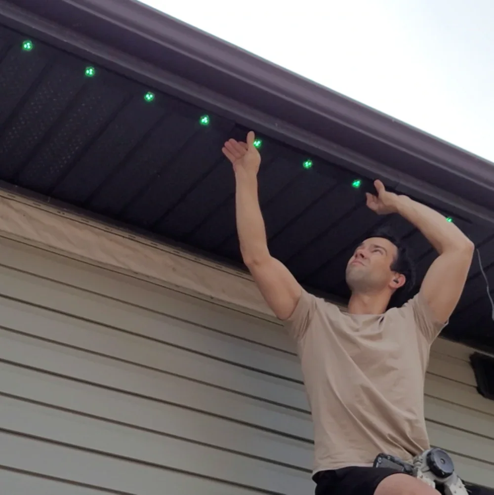 A man installing green LED lights underneath the eaves of a house.