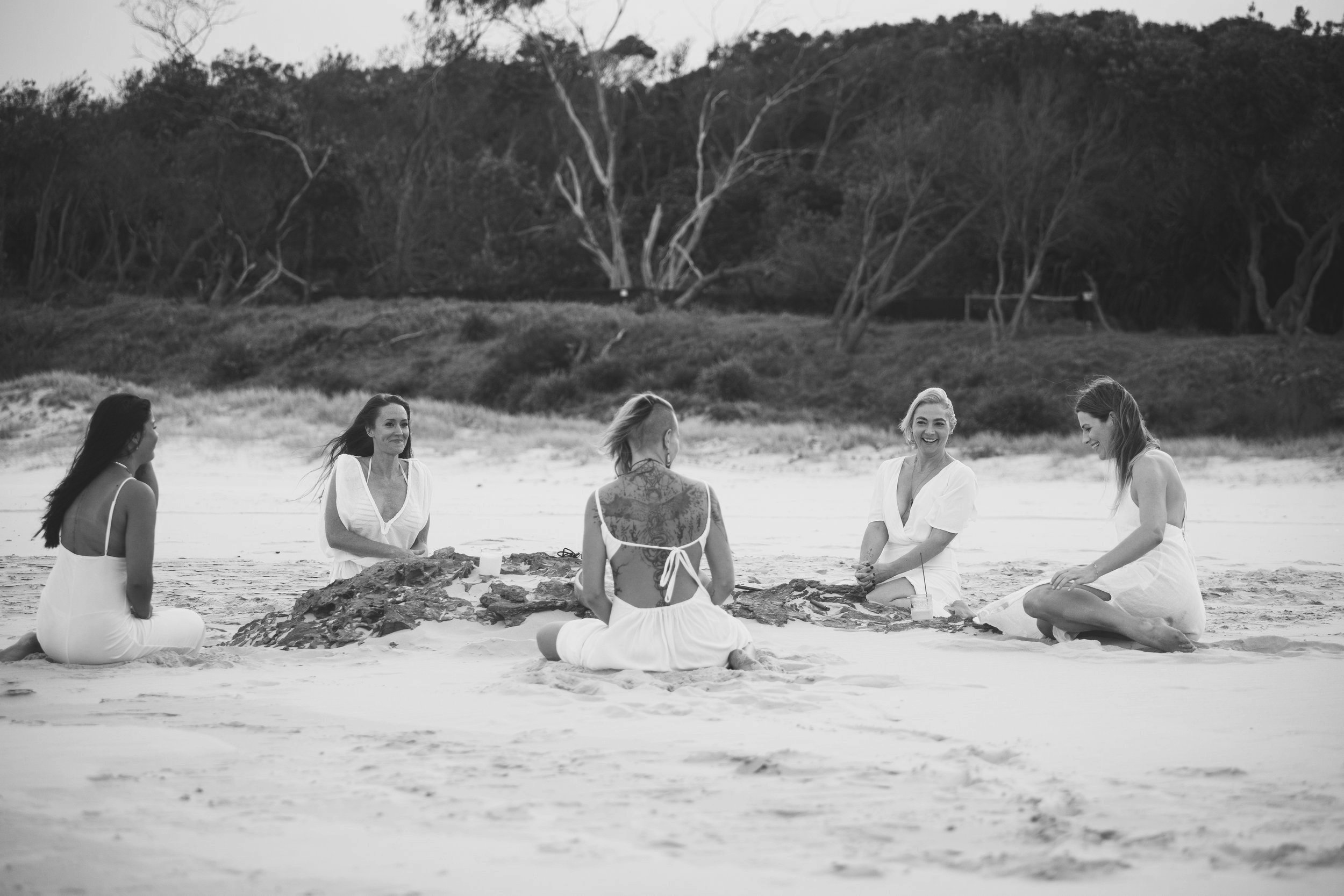 Six women dressed in white, sitting on the beach around a driftwood arrangement, smiling and engaging in conversation, with a backdrop of trees and sand dunes.