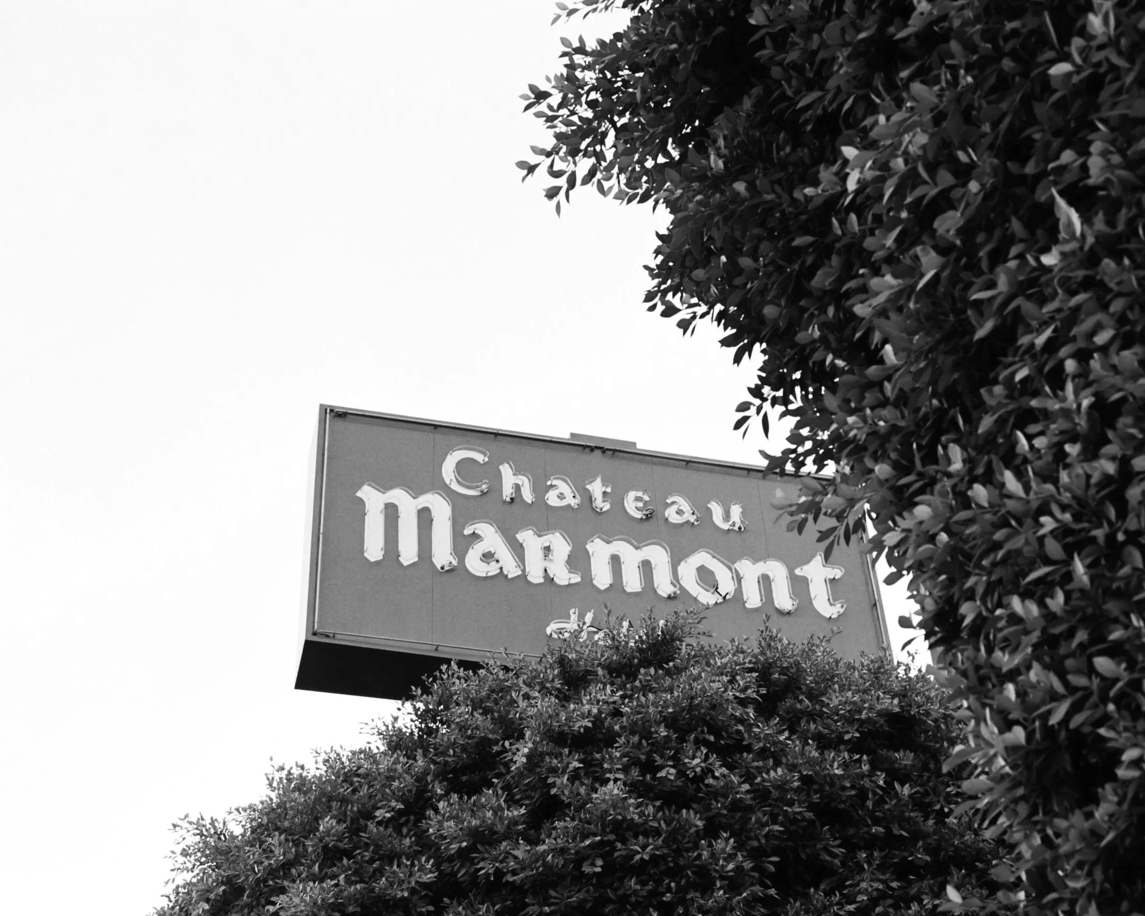 A black and white photo of a sign for Chateau Marmont hotel, partially obscured by trees.