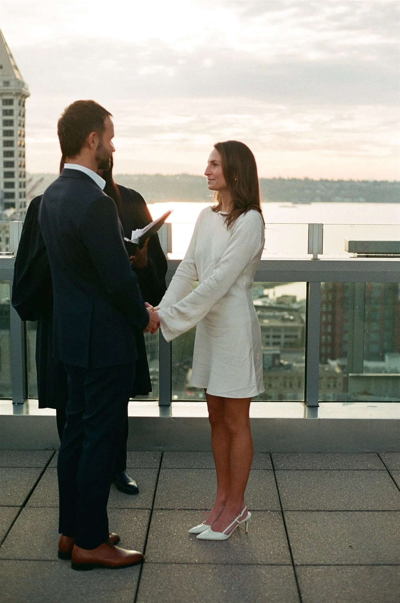 Couple holding hands during an intimate rooftop wedding ceremony at the Seattle Municipal Courthouse, with the city skyline and Elliott Bay visible behind them.