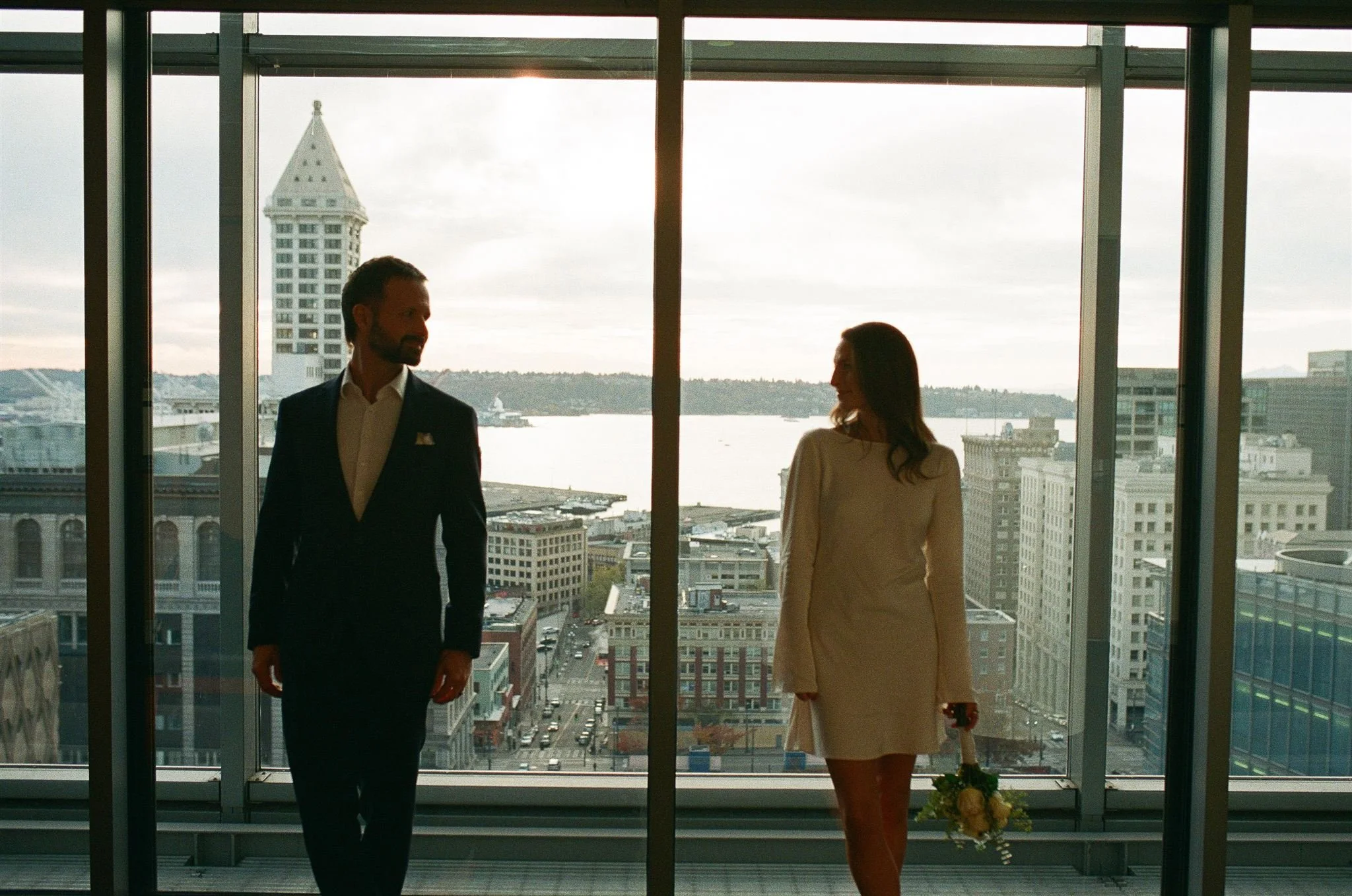 A couple walking side by side in front of tall windows overlooking downtown Seattle and Elliott Bay, the groom in a dark suit and the bride in a minimalist white dress holding a bouquet — a modern, rooftop courthouse wedding moment.