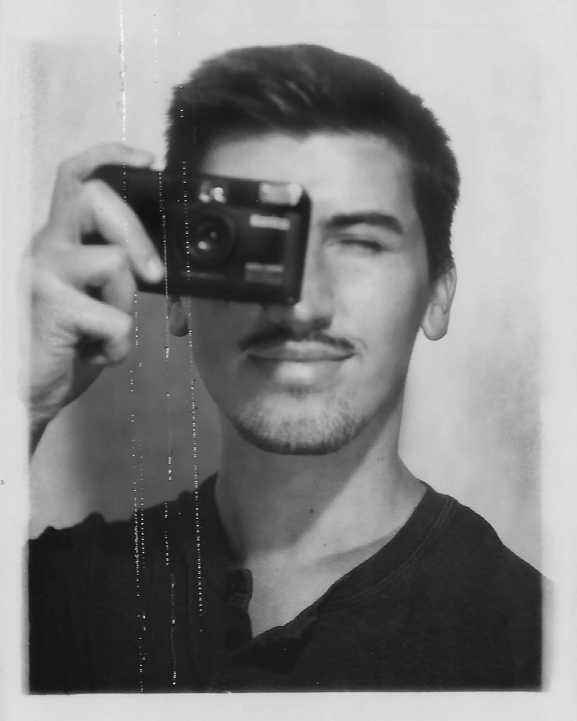 A black and white photo of a man with a mustache and goatee, smiling while taking a selfie with a camera, with one eye closed.