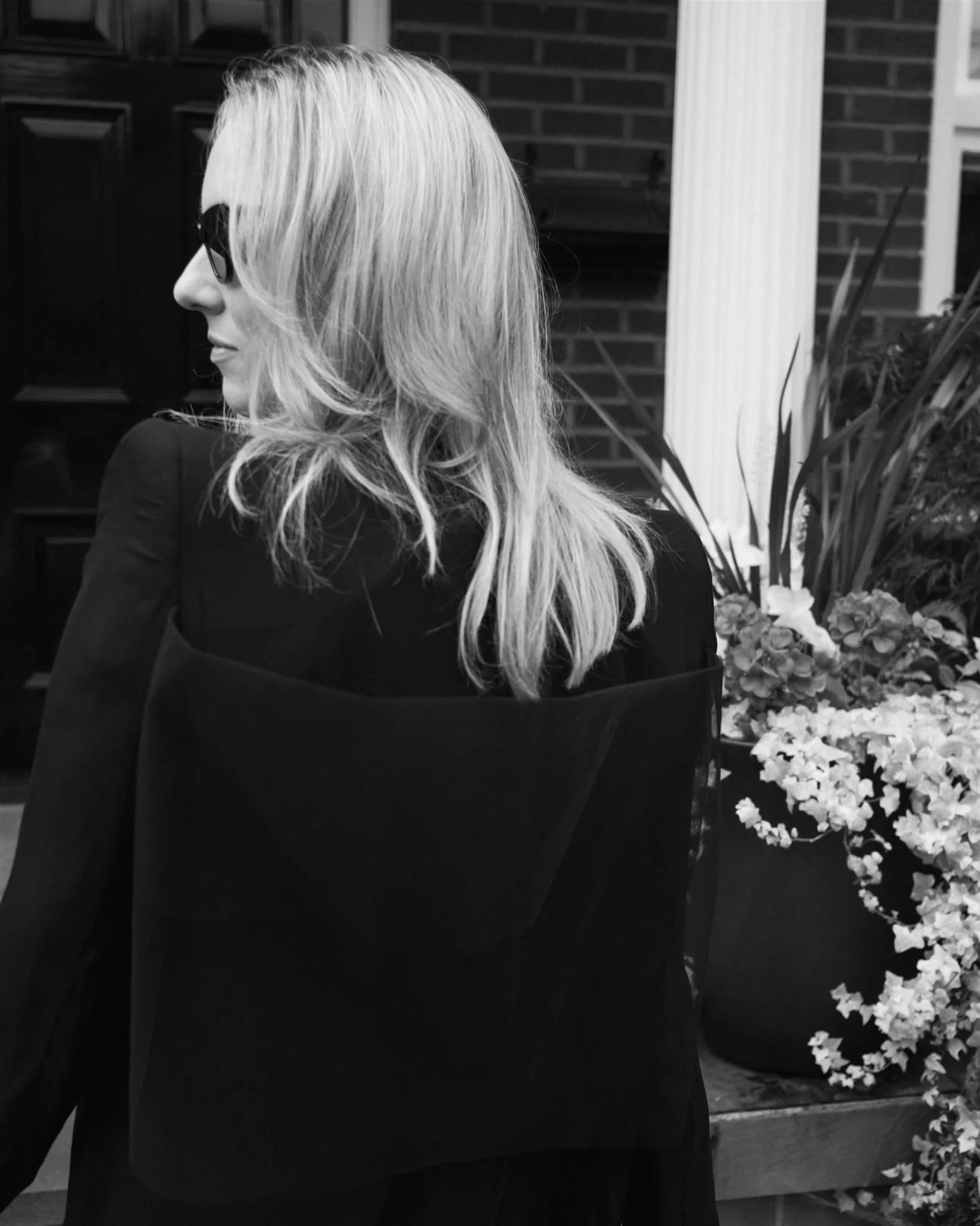 A woman with blonde hair wearing dark sunglasses and a dark outfit, sitting outdoors near a brick wall with plants and flowers nearby.
