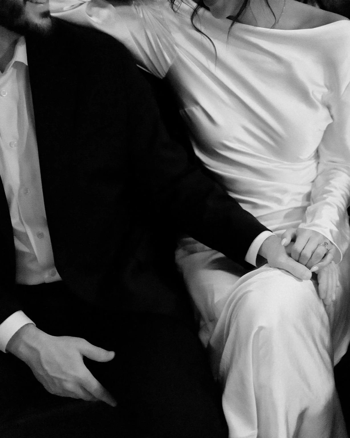 A black and white photo of a couple sitting close together, holding hands, with the woman's hand resting on her knee, both dressed in formal attire.