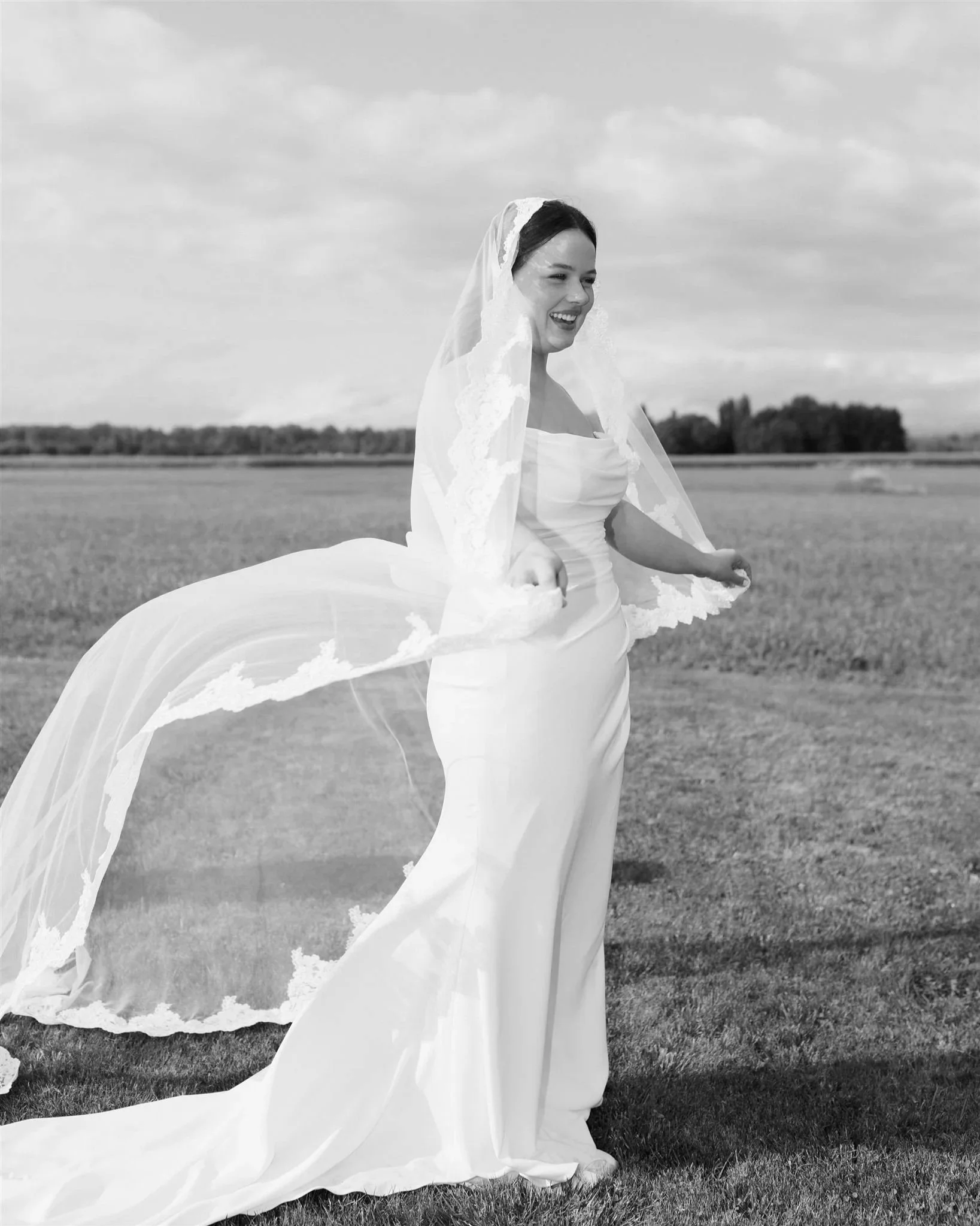 A smiling bride in a wedding dress standing in a field, holding her veil.