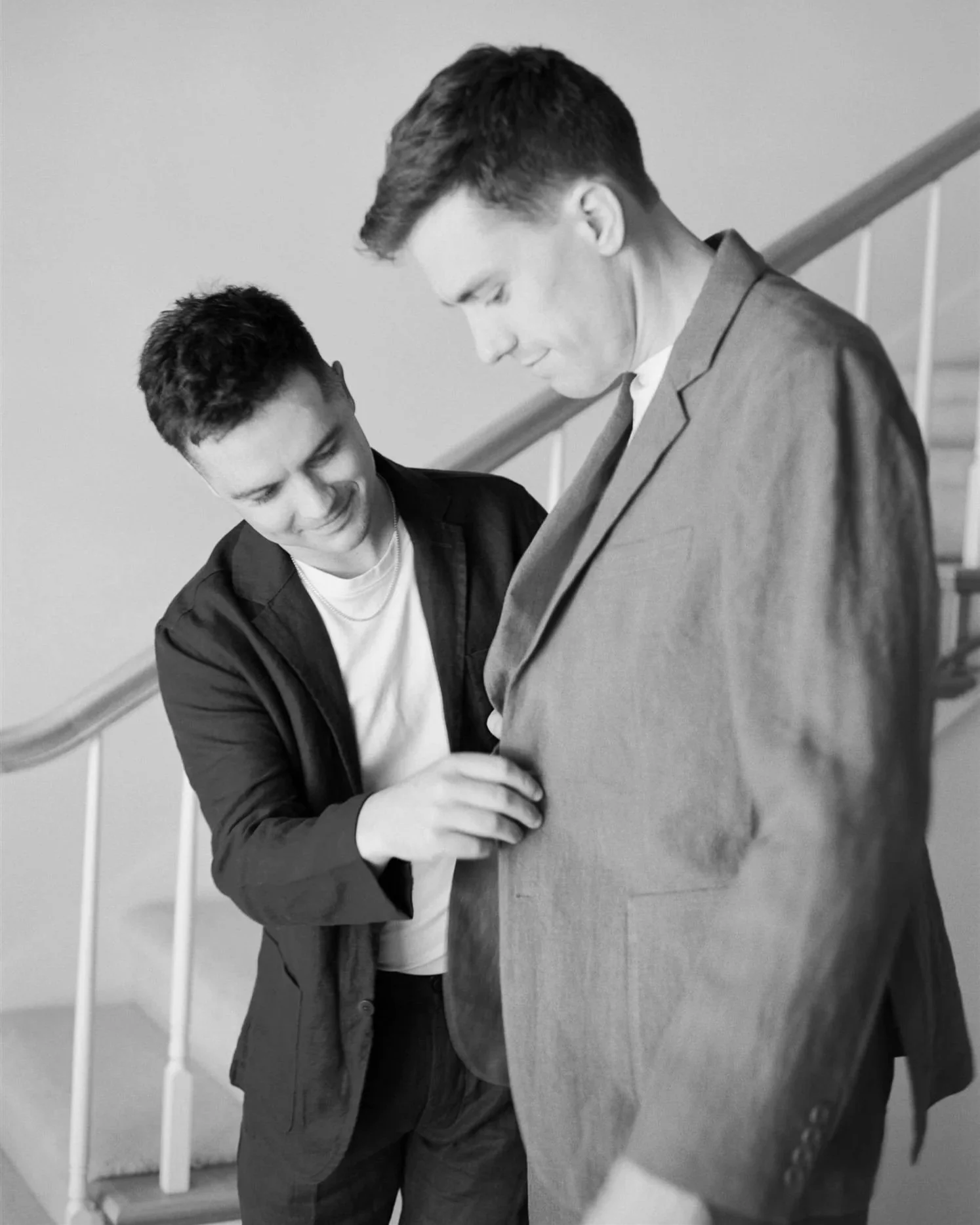 Two young men standing on a staircase, one adjusting the other's suit jacket, both smiling softly.