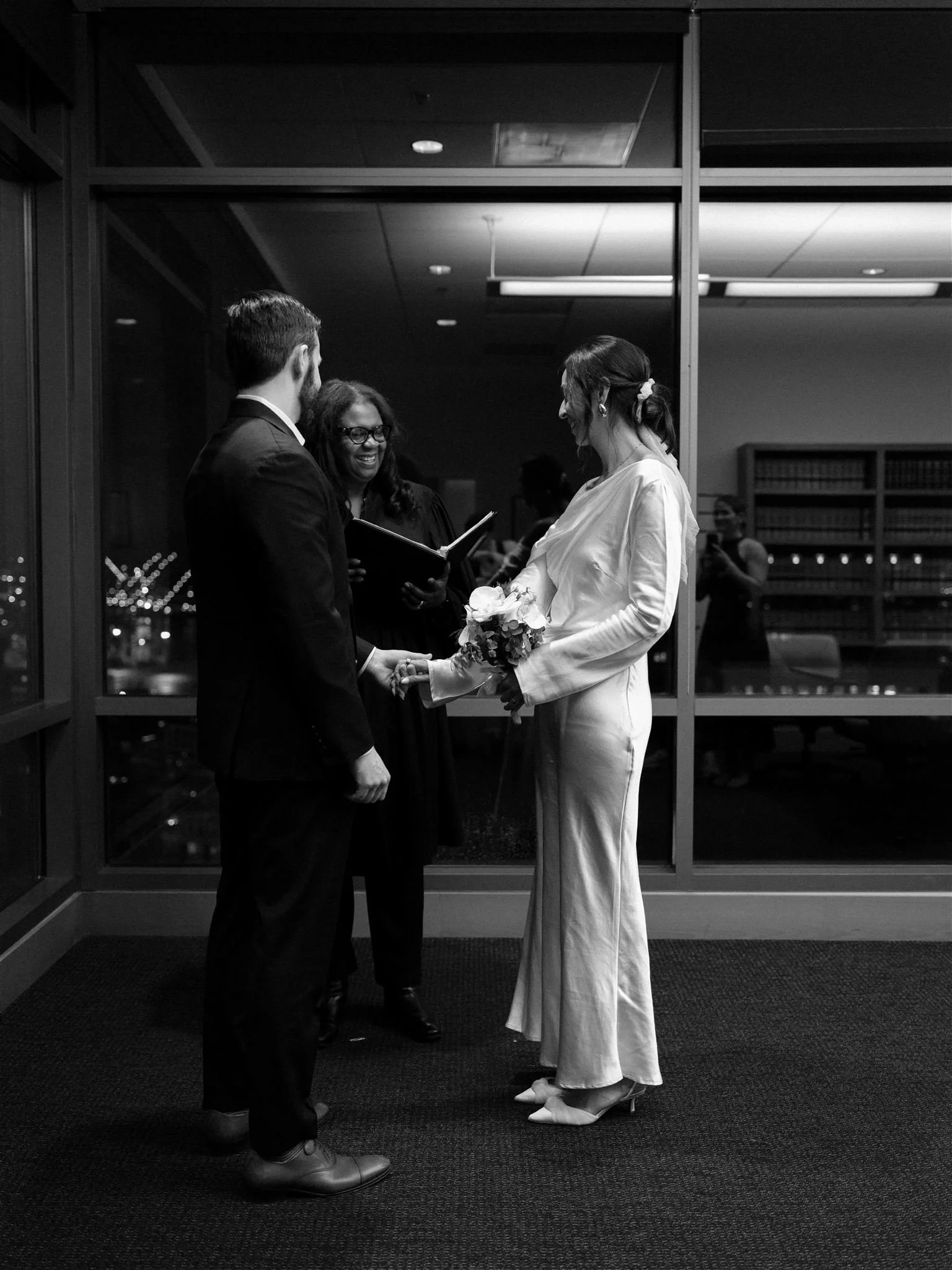 Couple holding hands during a courthouse wedding ceremony inside the Seattle Municipal Courthouse, with city lights visible through the windows in a black and white photograph.
