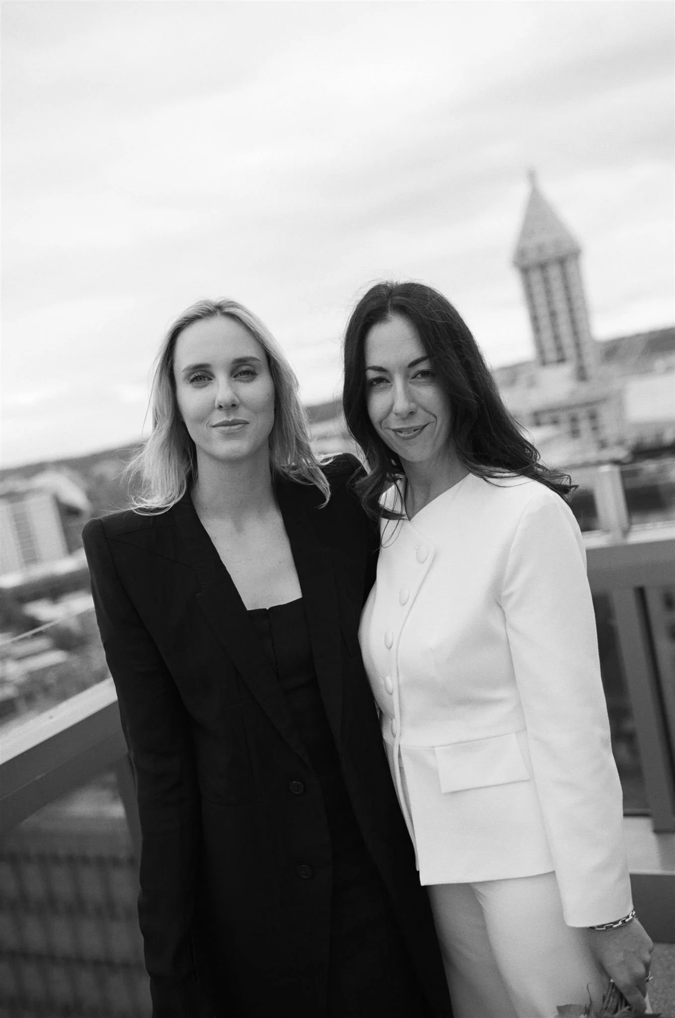Same-sex couple standing together on the rooftop of the Seattle Municipal Courthouse, dressed in modern black and white suits with the Seattle skyline softly blurred behind them.