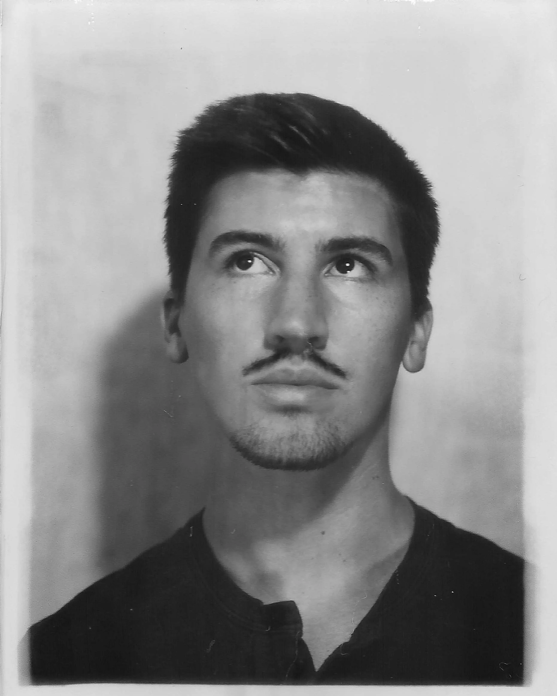 Black and white portrait of a young man with dark hair, a light mustache, and goatee, looking slightly upward and to the side.