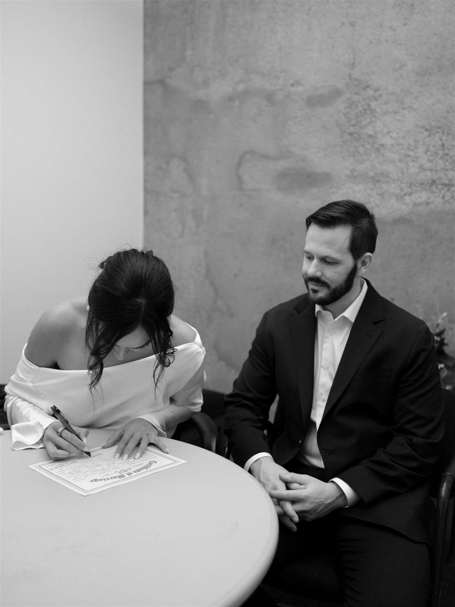 Bride signing the marriage license inside the Seattle Municipal Courthouse while her partner sits beside her, photographed in black and white during an intimate courthouse wedding.