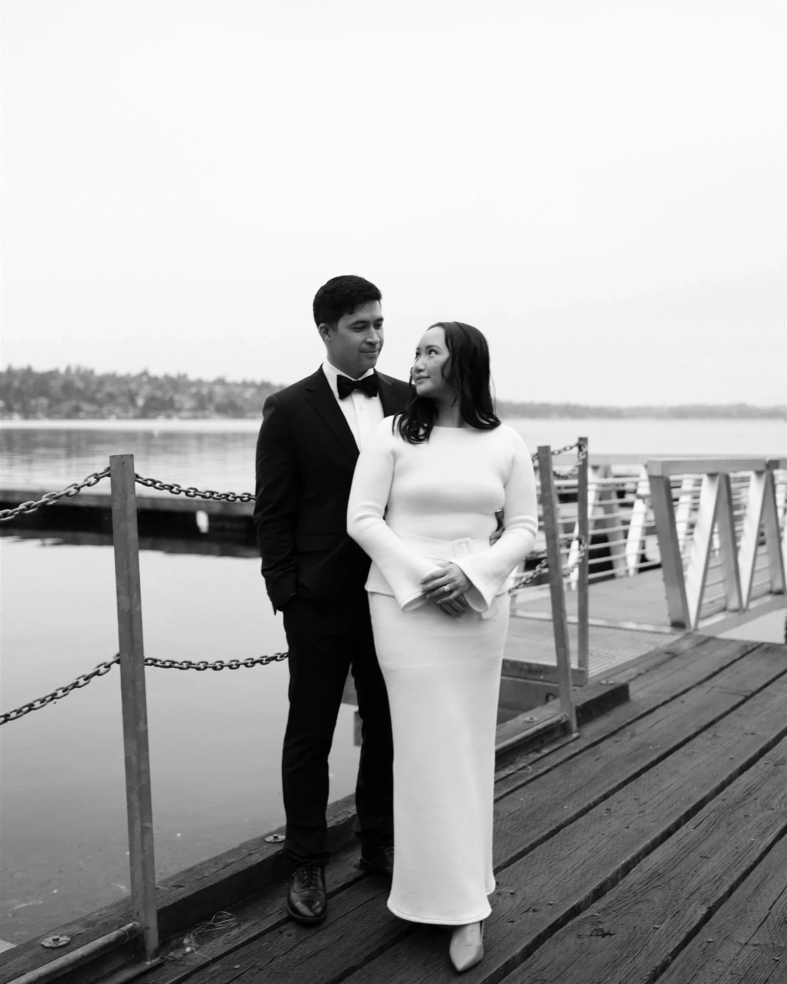 A black and white photo of a couple standing on a wooden dock by a lake, dressed formally. The man is wearing a tuxedo with a bow tie, and the woman is in a long, elegant dress. They are looking at each other lovingly.