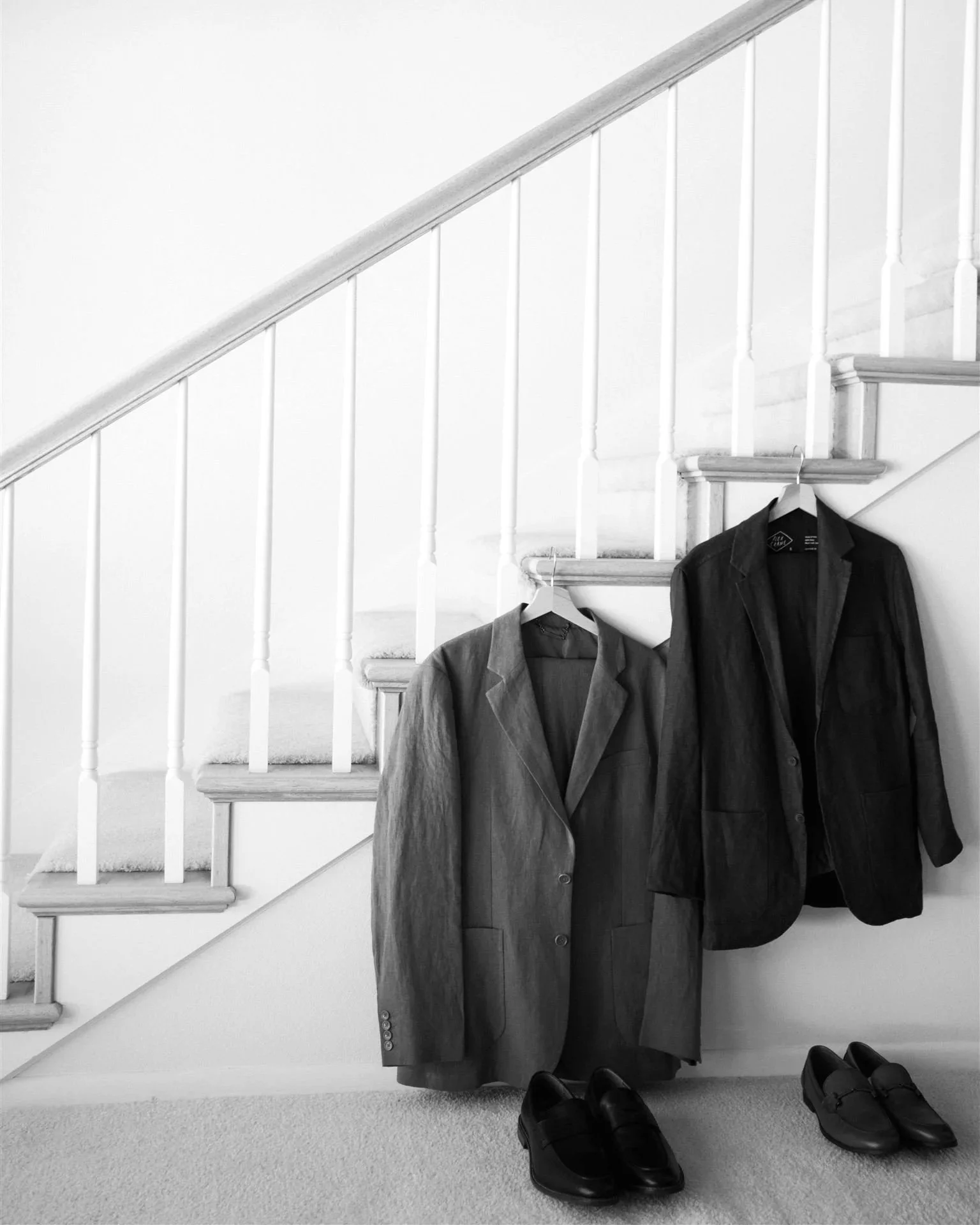 Two suits hanging on stair railing with dress shoes underneath in a black-and-white photo.