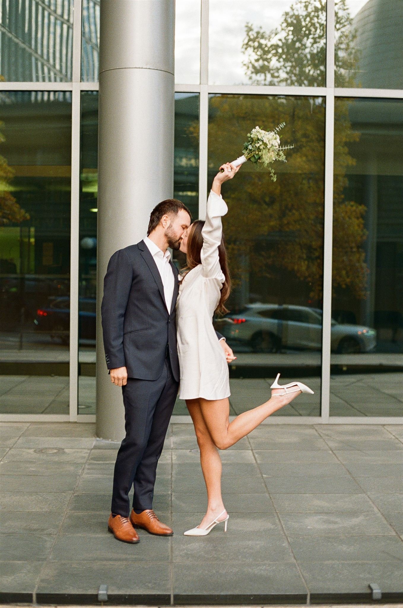 Couple kissing outside the Seattle Municipal Courthouse, the bride lifting her bouquet and leg in celebration while wearing a minimalist white dress and heels