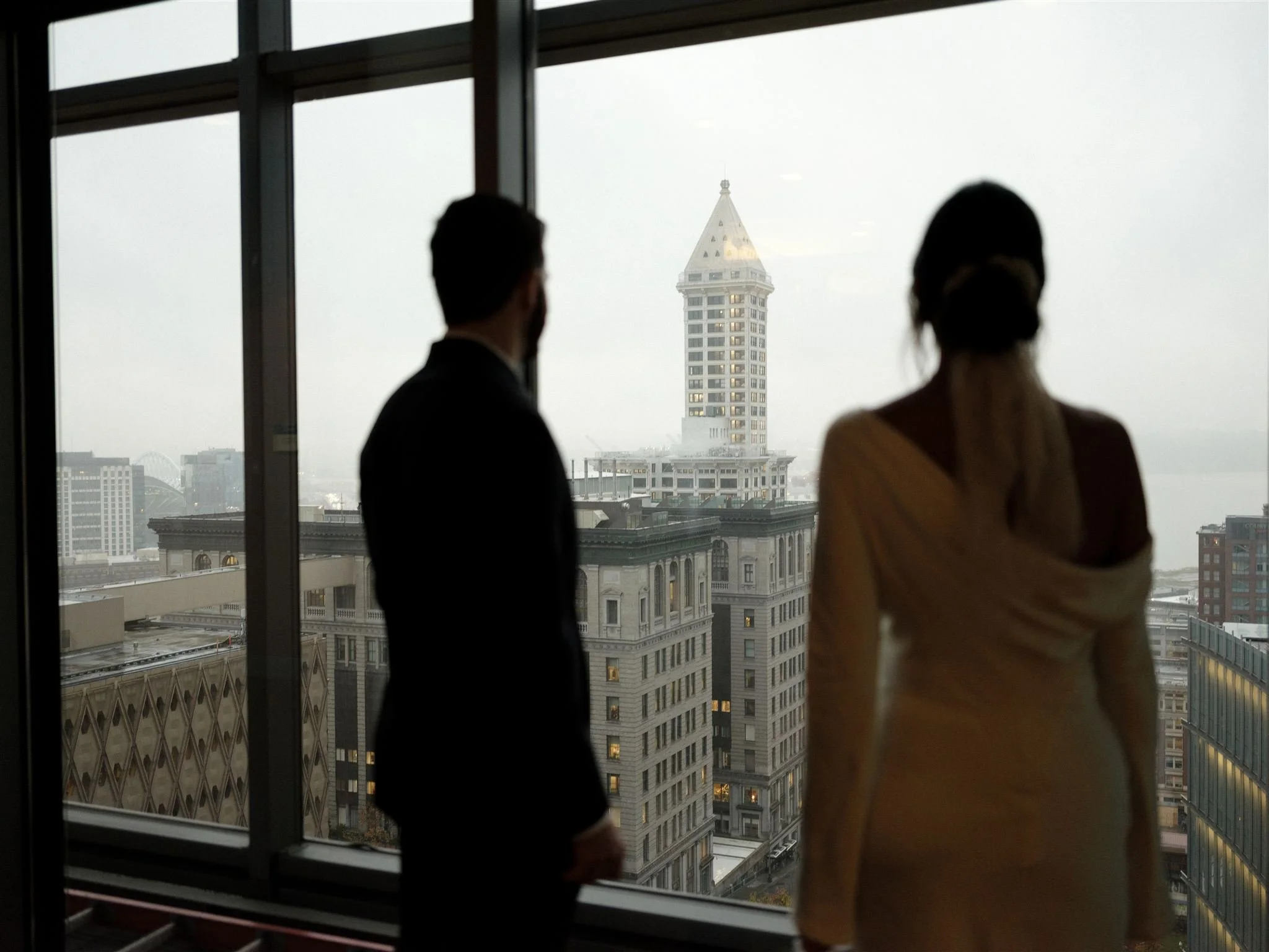 Couple standing by floor-to-ceiling windows inside the Seattle Municipal Courthouse, looking out over downtown Seattle and the Smith Tower on an overcast day.