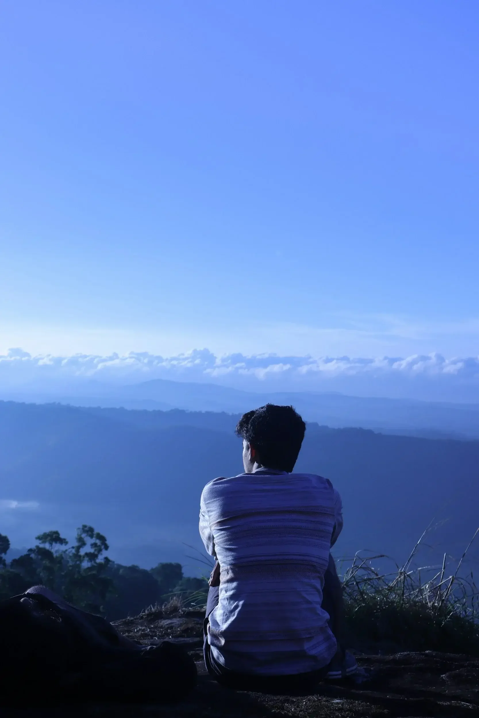 Person with short curly hair wearing a gray and white striped shirt sitting on rocky ground, looking out over mountains and a valley with a clear blue sky and clouds in the background before relationship therapy.