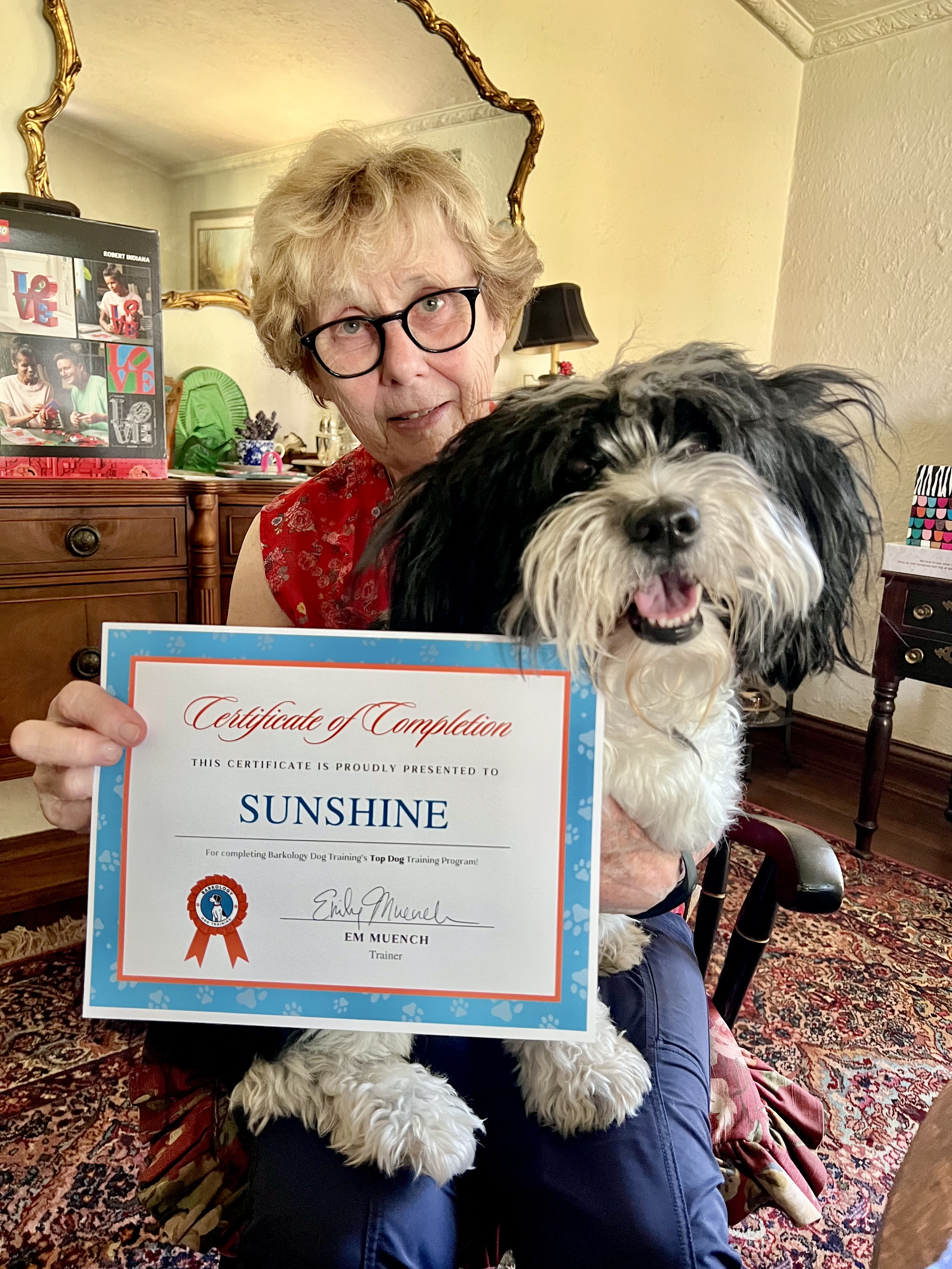 dog parent holds dog trainer graduation diploma with her small black and white doodle sitting on her lap and they are both smiling
