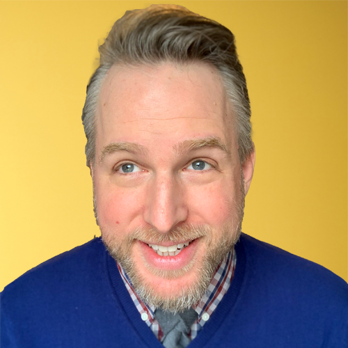 A man with a beard smiling and looking to the side against a yellow background, wearing a blue sweater over a plaid shirt.