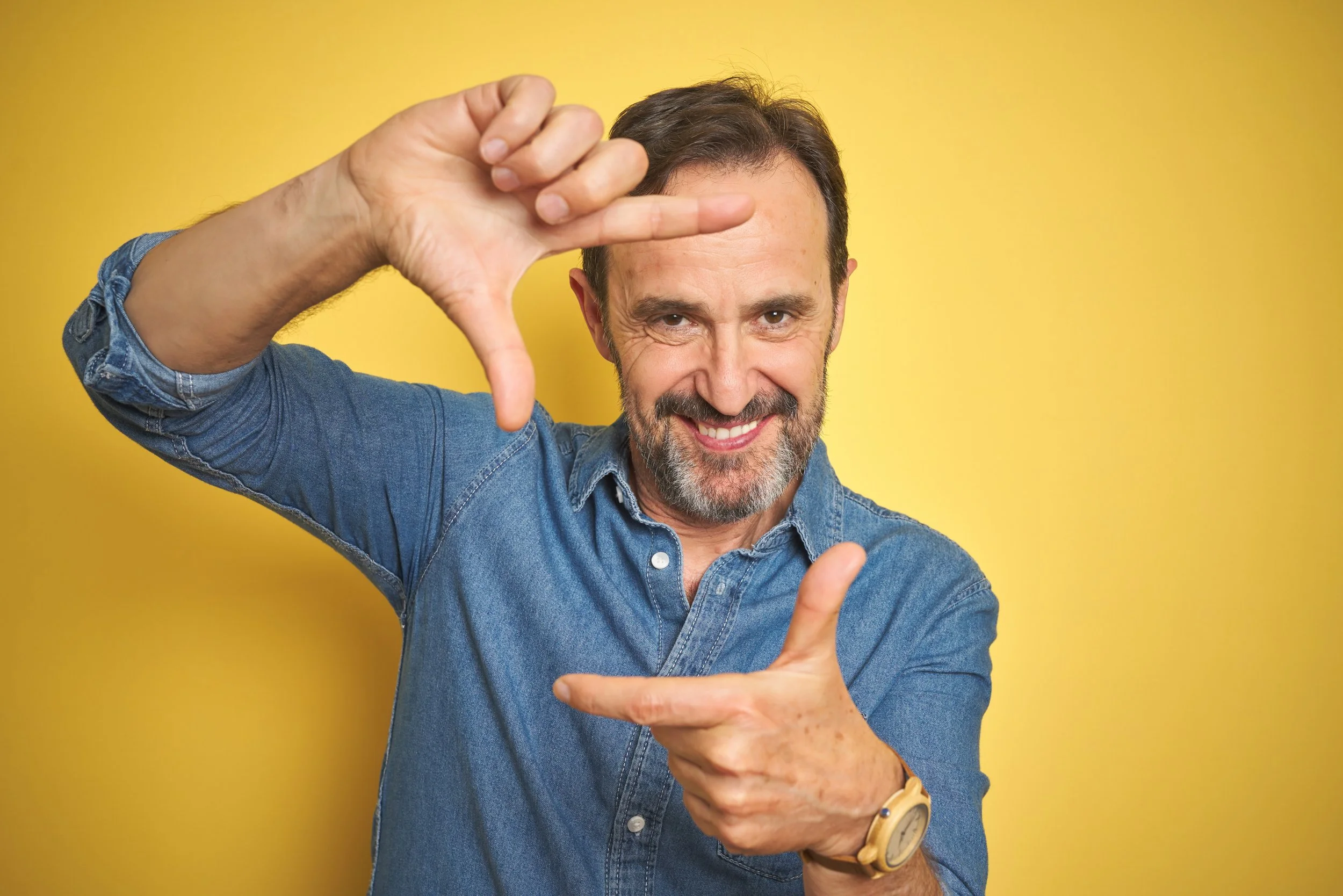 A middle-aged man with a beard and short brown hair, wearing a denim shirt and a brown watch, smiling and making a frame with his hands in front of a yellow background.